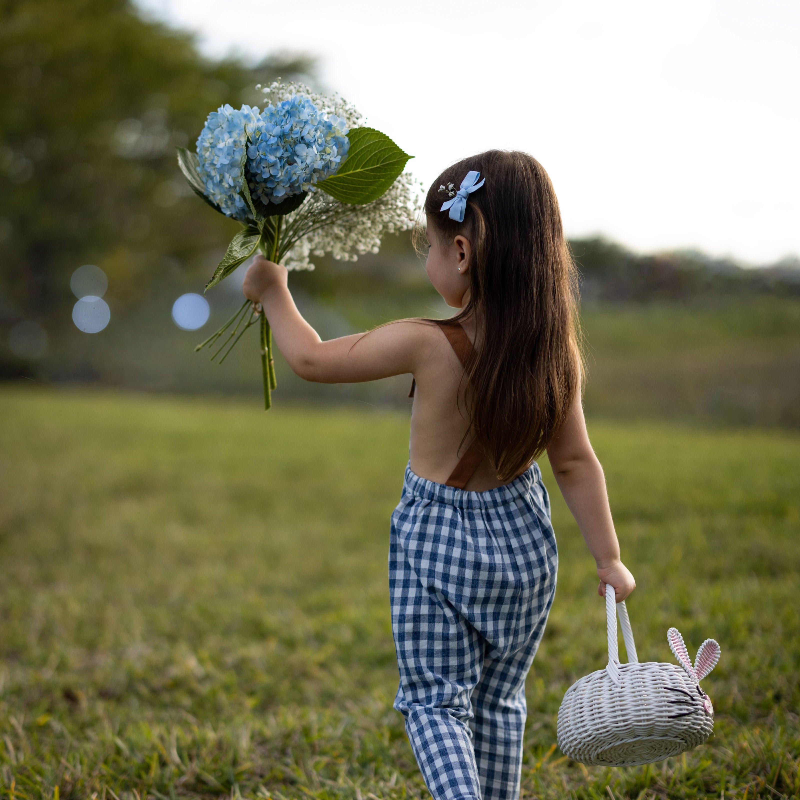 Blue Gingham Linen Long Overalls on a little girl from the back during Easter, showcasing the gender-neutral design perfect for passing down, breathable fabric for all-day comfort, stylish pleats for elegance, durable leather-looking straps, and adjustable straps for a perfect fit. Made by a sustainable brand committed to eco-friendly practices, promoting responsible fashion for children.