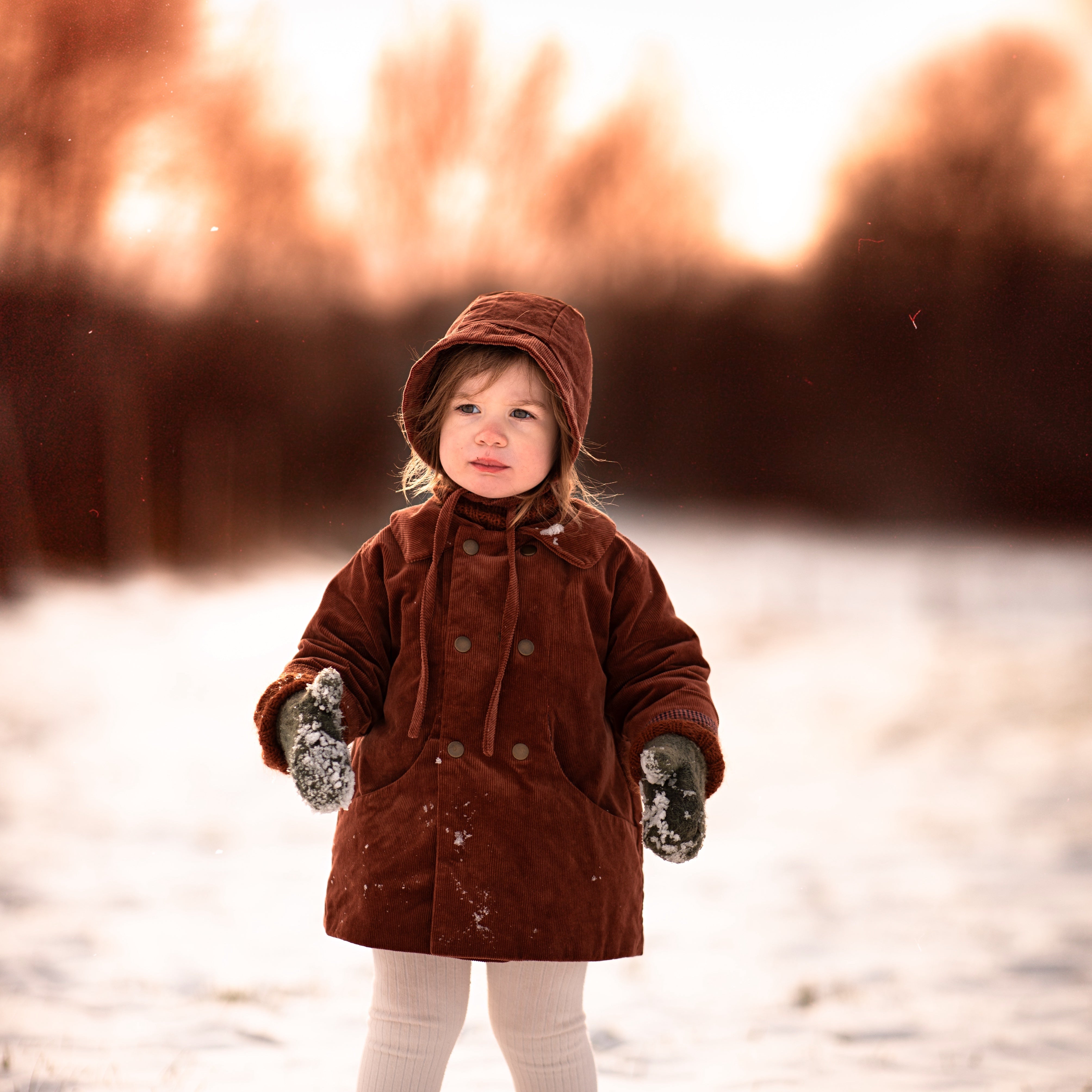 Little girl in the snow in a rust coat and matching hat with blurry background