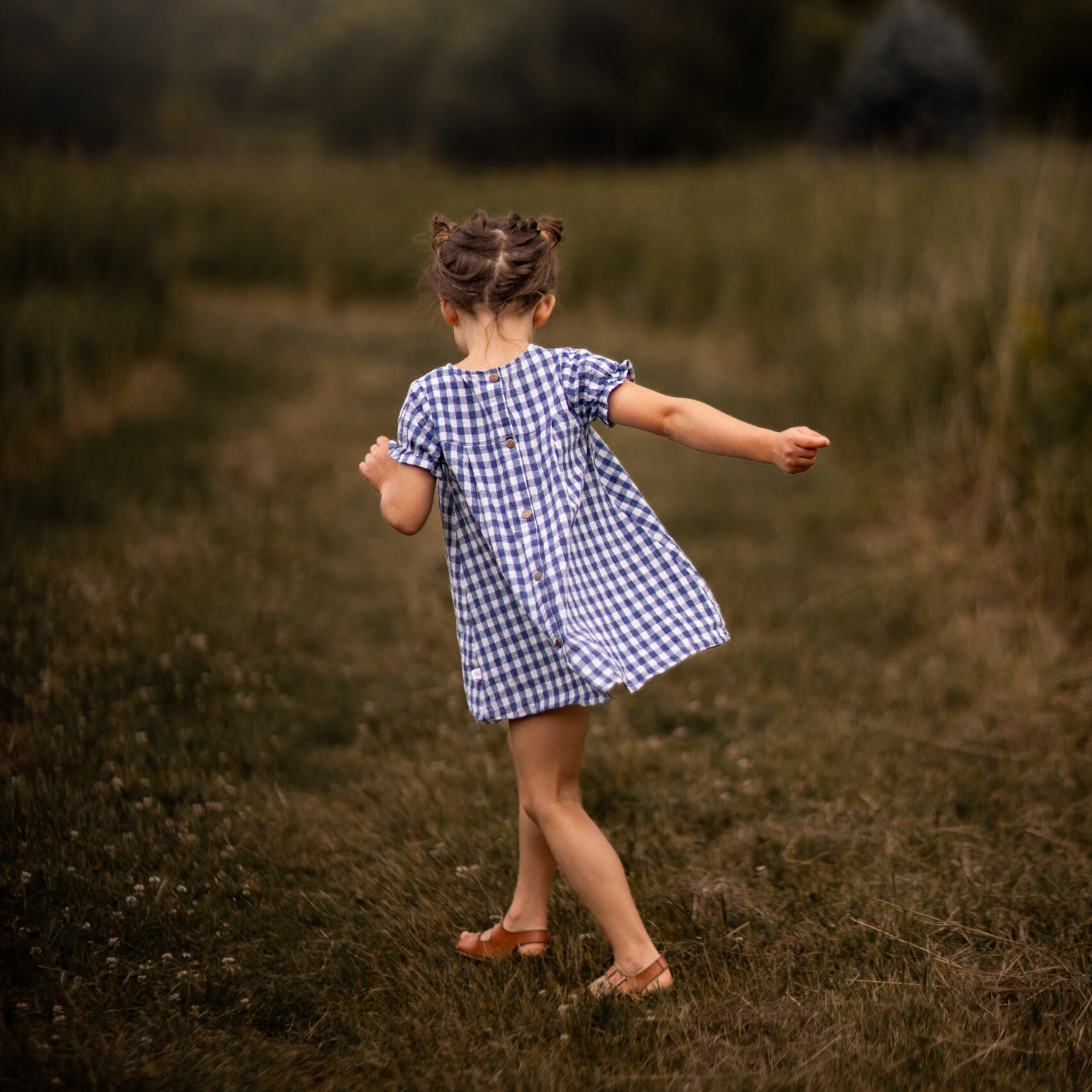 A little girl stands with her back to the camera, showcasing the Blue Gingham linen dress. The dress features a pin-tucked front and a yoke with two pleats at the back, emphasizing its charming design. The delicate ruffles at the short sleeve hem add a sweet touch, while the A-line shape allows for comfortable movement. Her hair is styled up, and she enjoys the sunny day in a picturesque field, highlighting the dress's practicality and style.