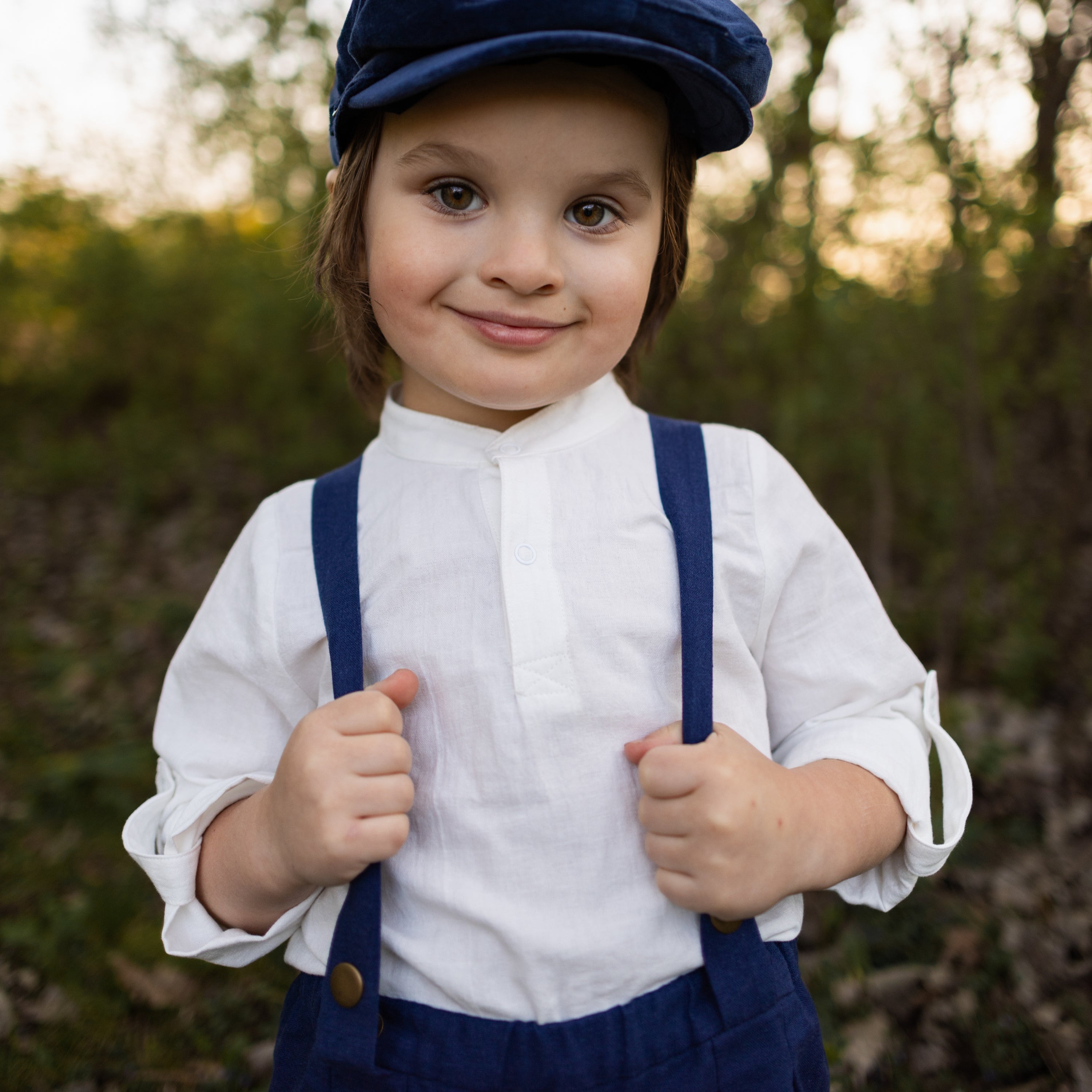 A close-up of boy, capturing his charming smirk and elegant features, highlighting the details of his outfit, including the navy linen suspender shorts and white Henley shirt, set against a soft background that enhances his stylish appearance.