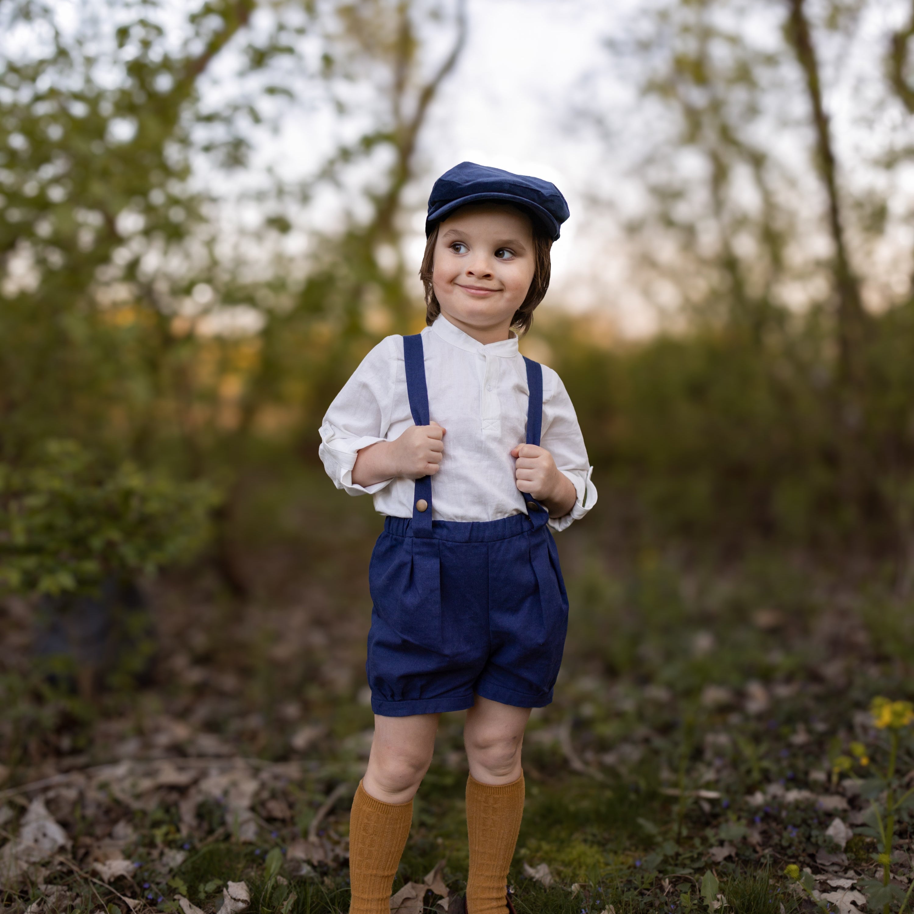 Child in vintage outfit with suspenders and cap standing in a forest
