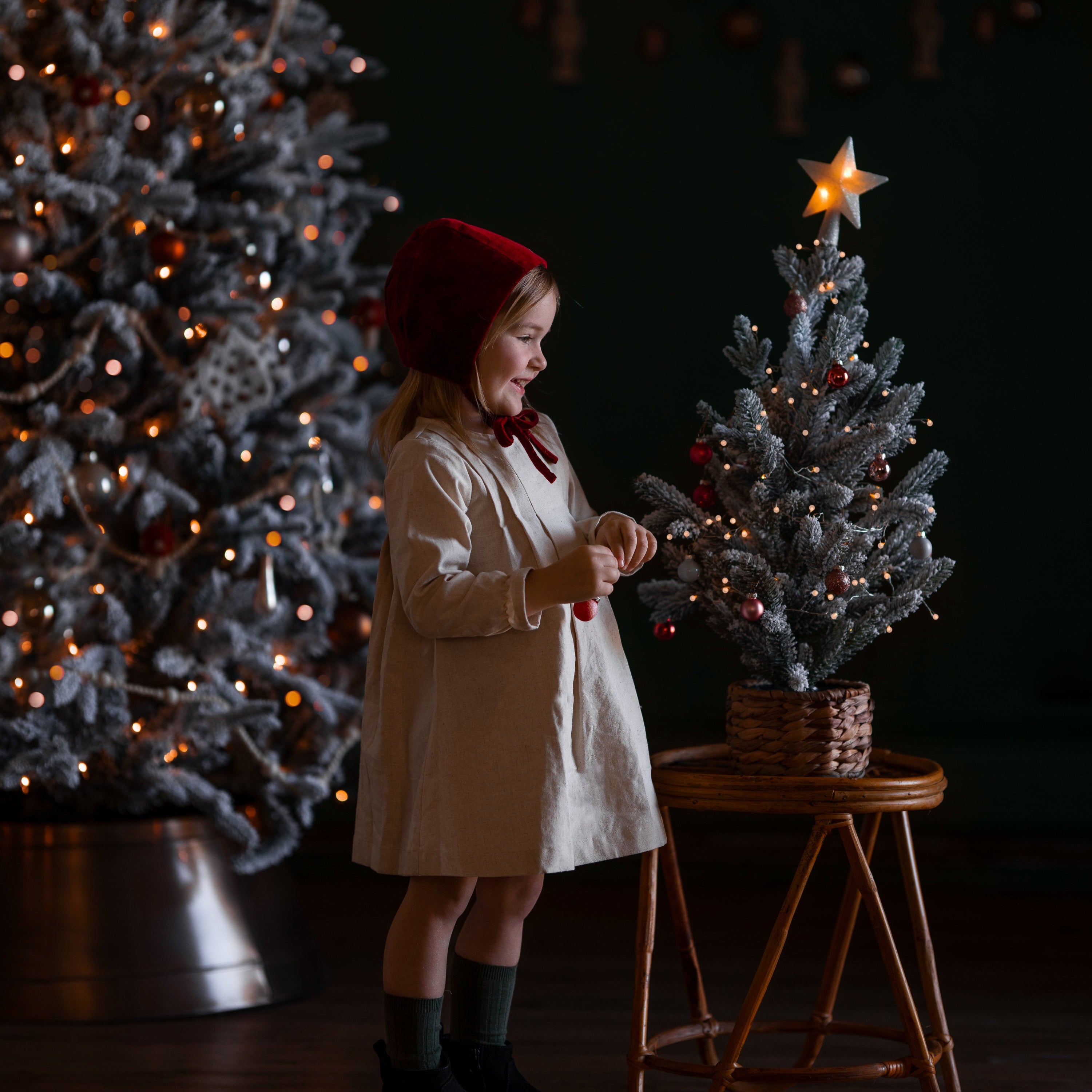 Child in a white dress and red hat standing next to a small Christmas tree with a star on top, in a dimly lit room.