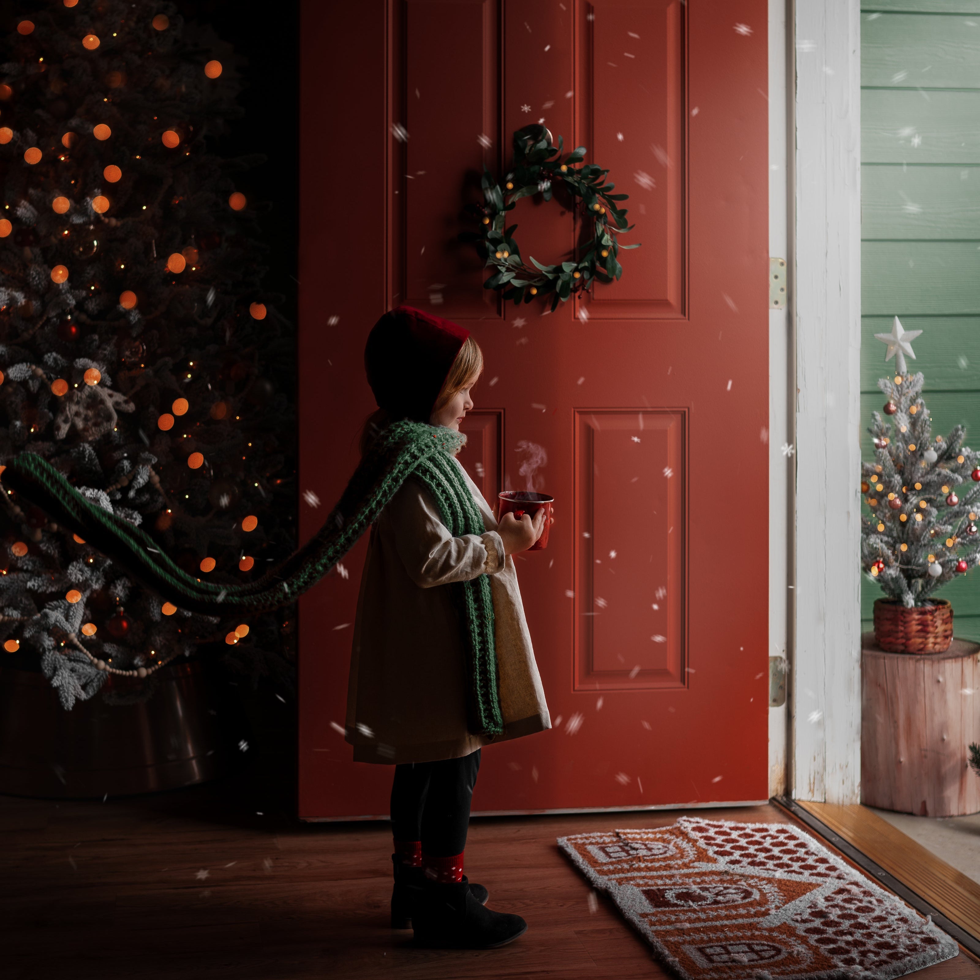 Girl standing in front of open door with snow flying in wearing a red velvet bonnet and a natural linen dress with a long green scarf