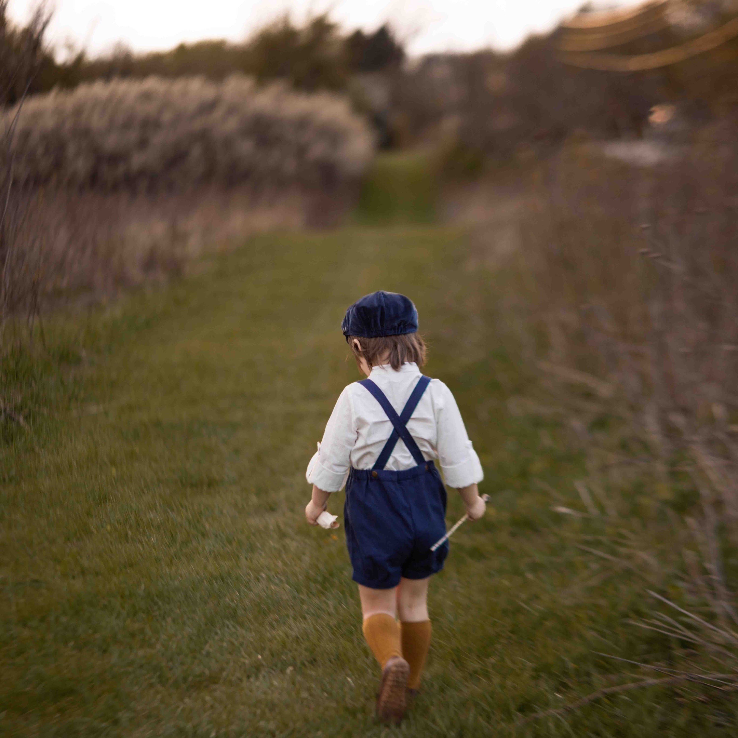 Child walking away in a field wearing a white shirt, blue overalls, and brown boots.