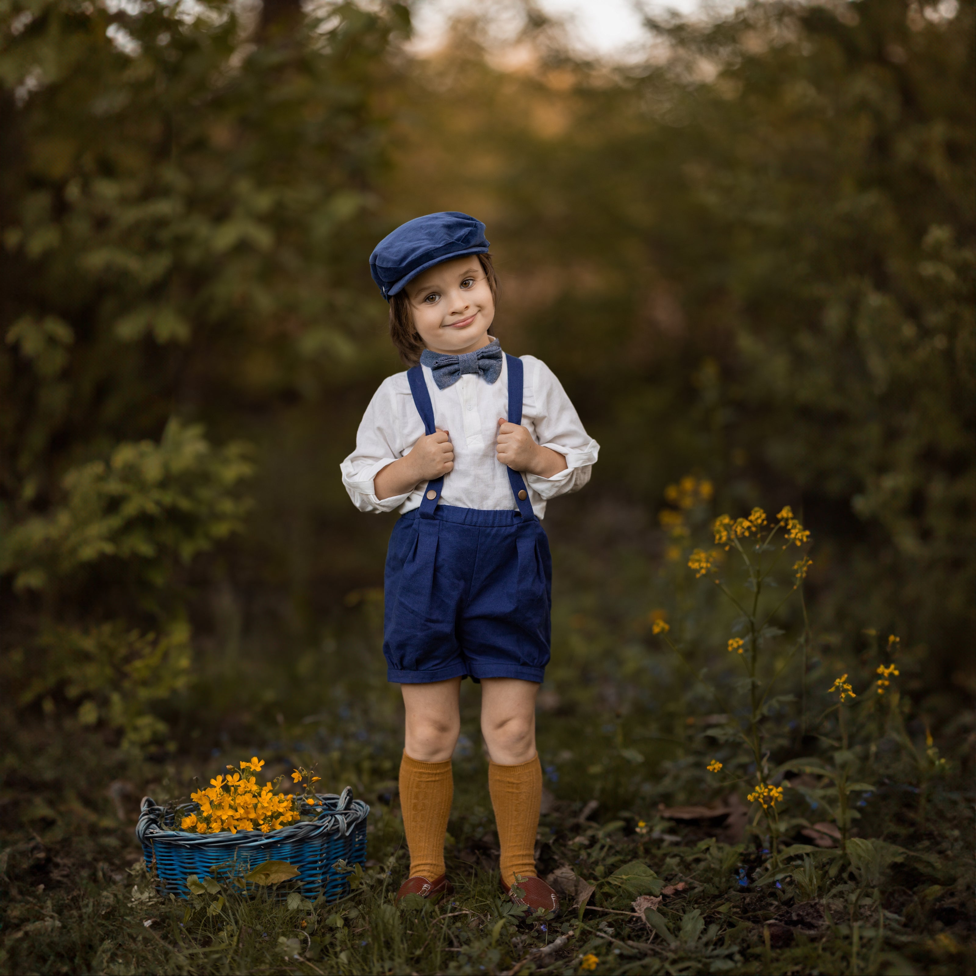 A model boy posing with a smirk, exuding elegance while wearing his navy linen suspender shorts and white Henley shirt. His confident expression and stylish outfit create a charming and sophisticated look.