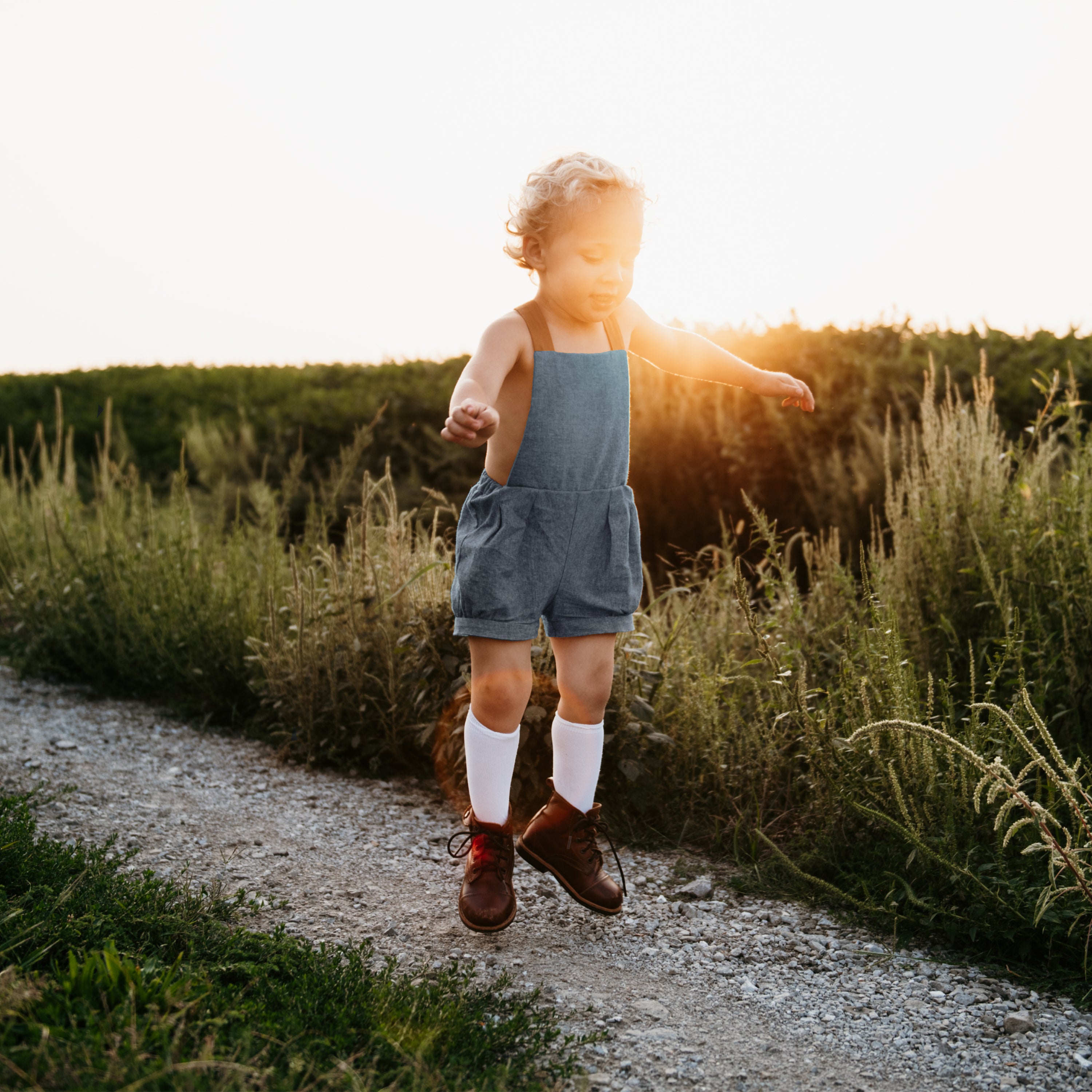 Energetic 3-year-old boy in chambray blue linen short overalls, captured mid-jump, demonstrating the freedom of movement and all-day comfort for active children