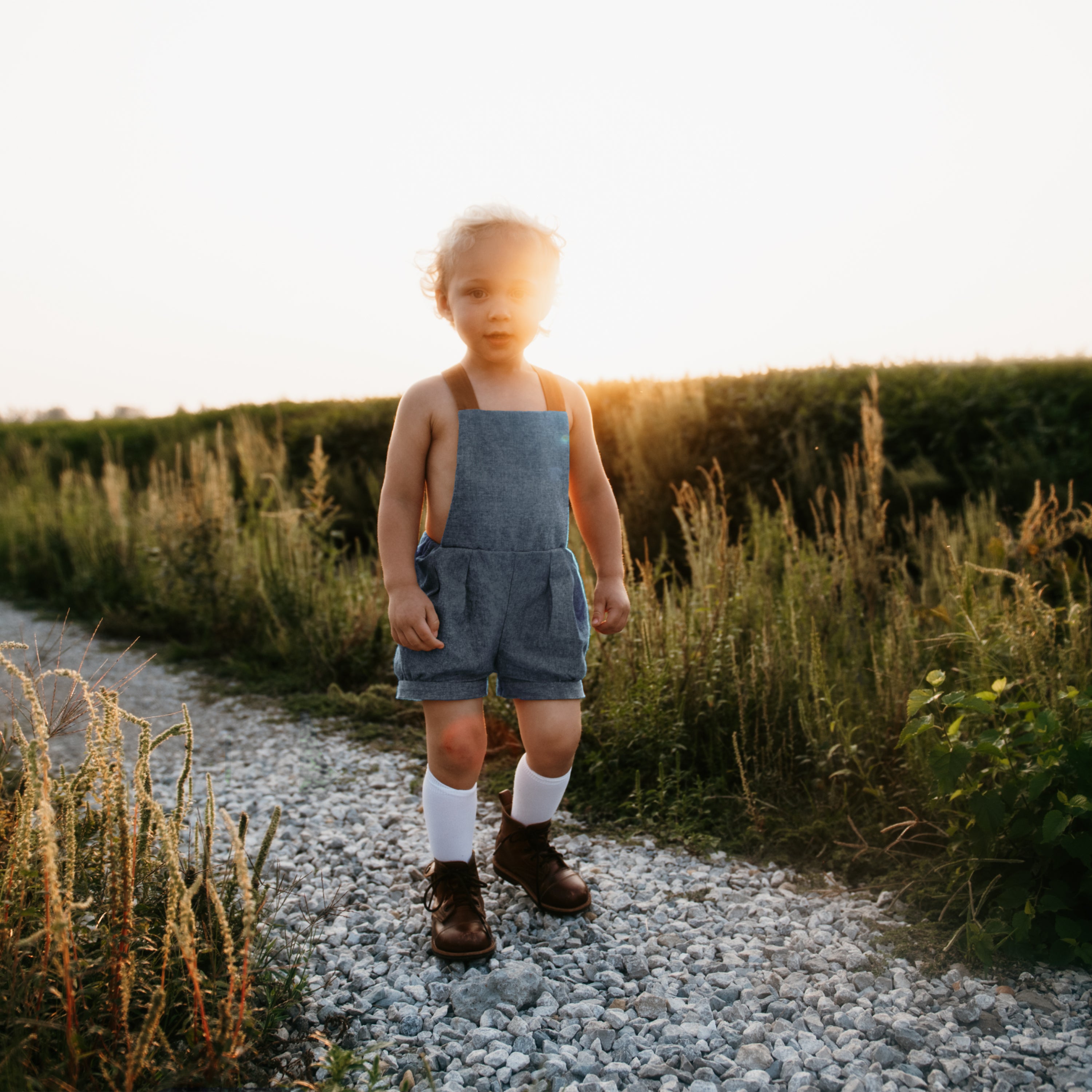 A joyful 3-year-old boy wearing chambray blue linen short overalls, playing in a sunlit field, highlighting the comfort and versatility of natural, sustainable clothing for kids.