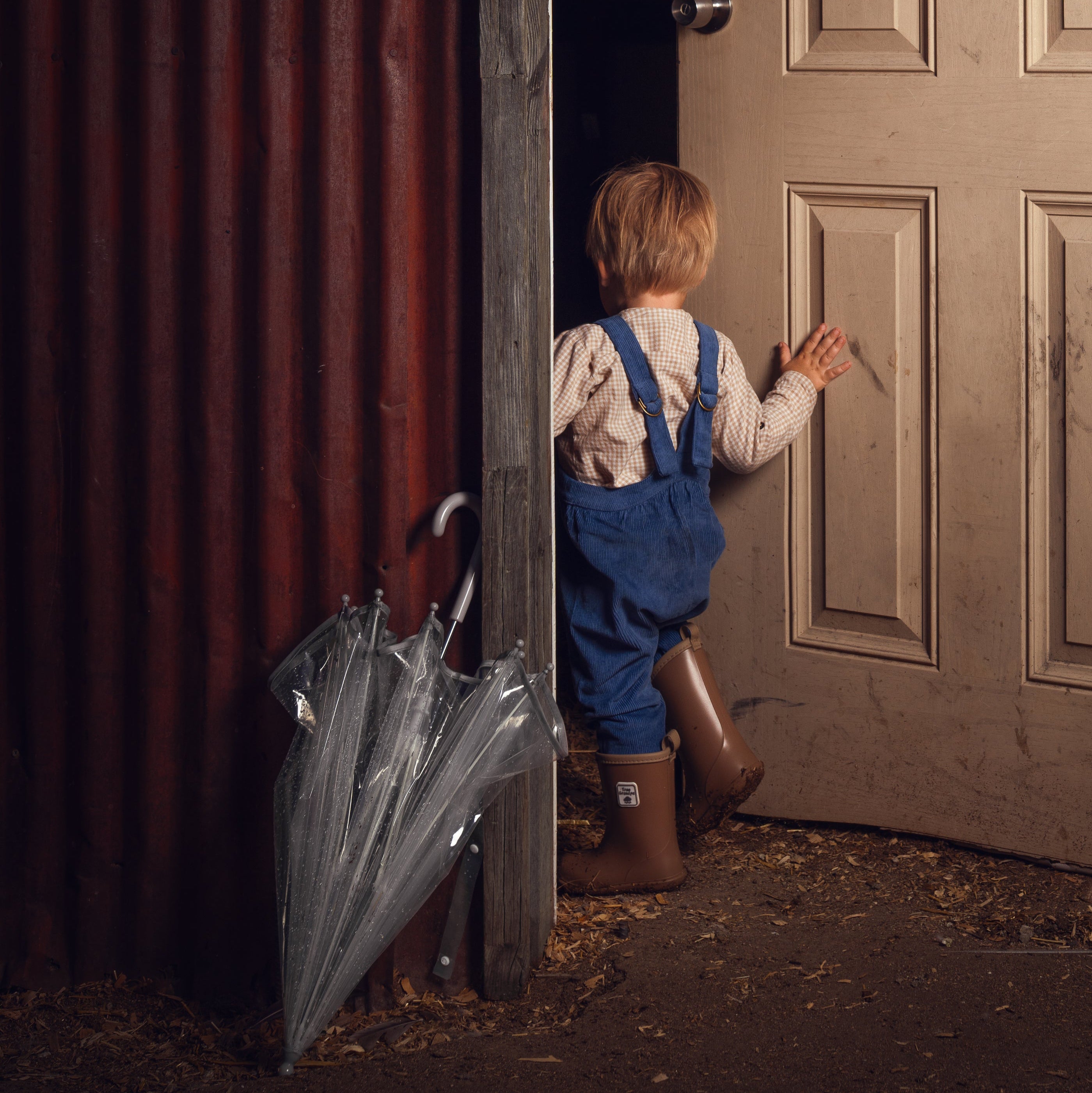 Child in blue overalls and rain boots standing in a doorway with an umbrella, looking out into a darkened room.