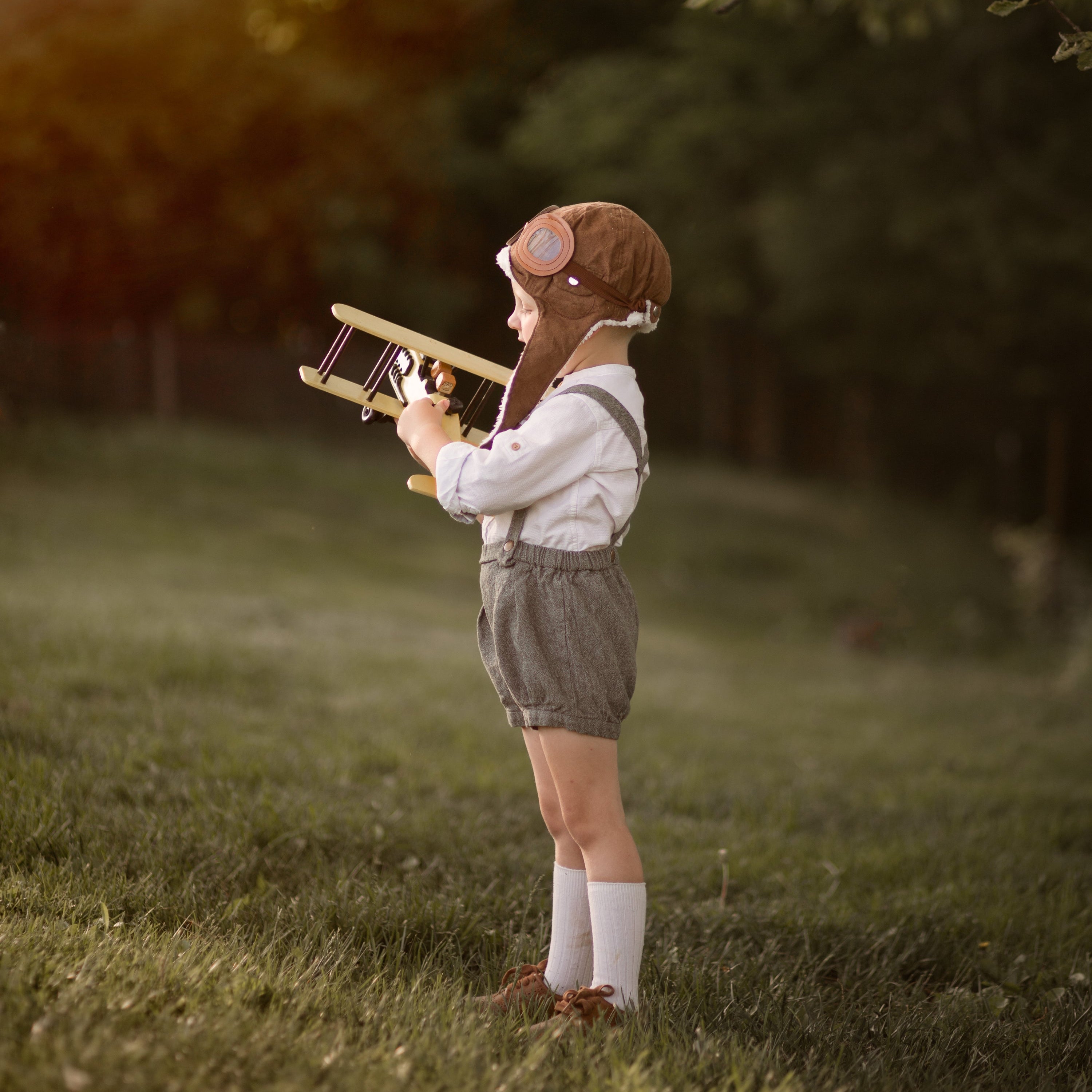 Child in green suspender shorts holding a toy airplane in a natural setting with trees and grass.
