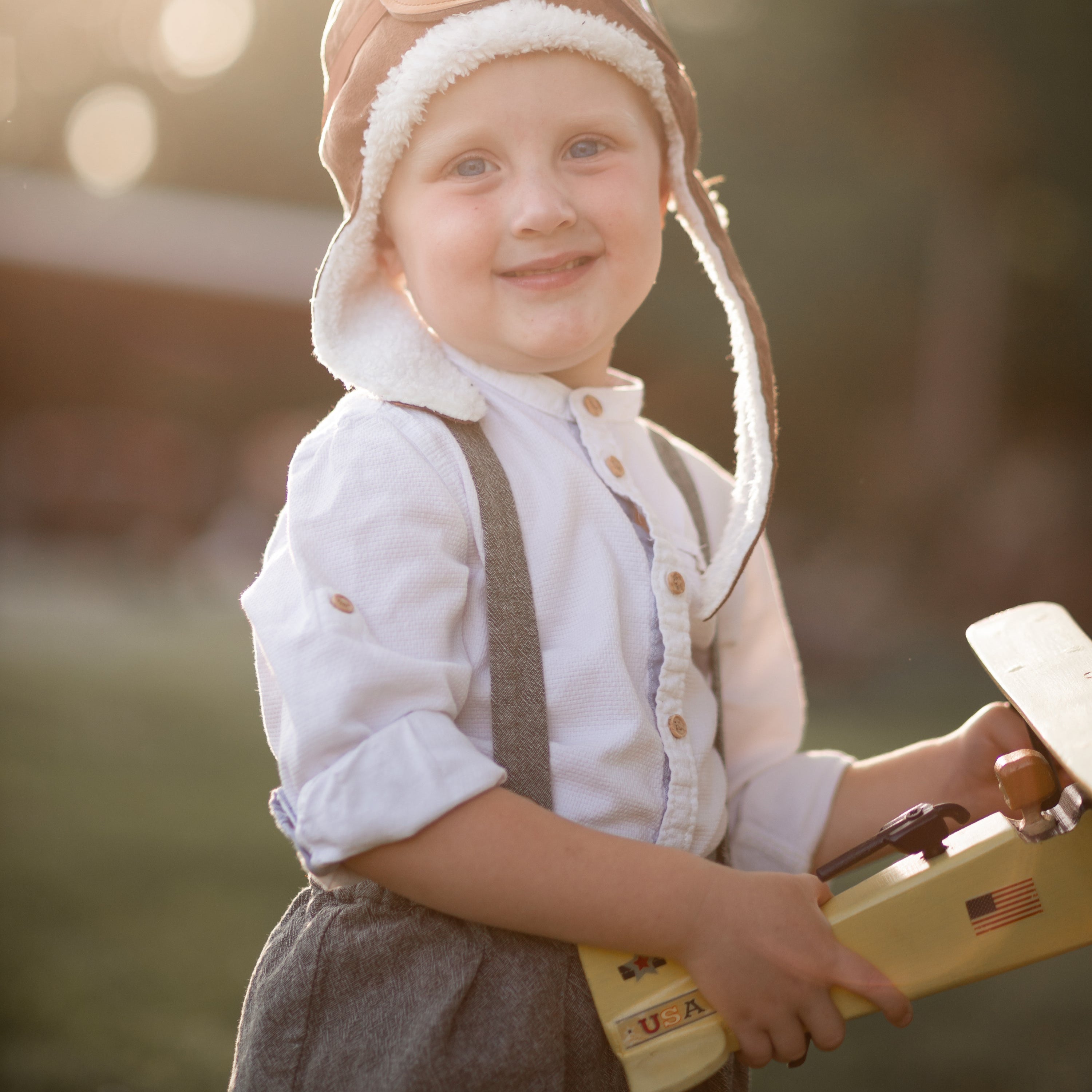 Child wearing a pilot hat and holding a toy airplane outdoors with a blurred background