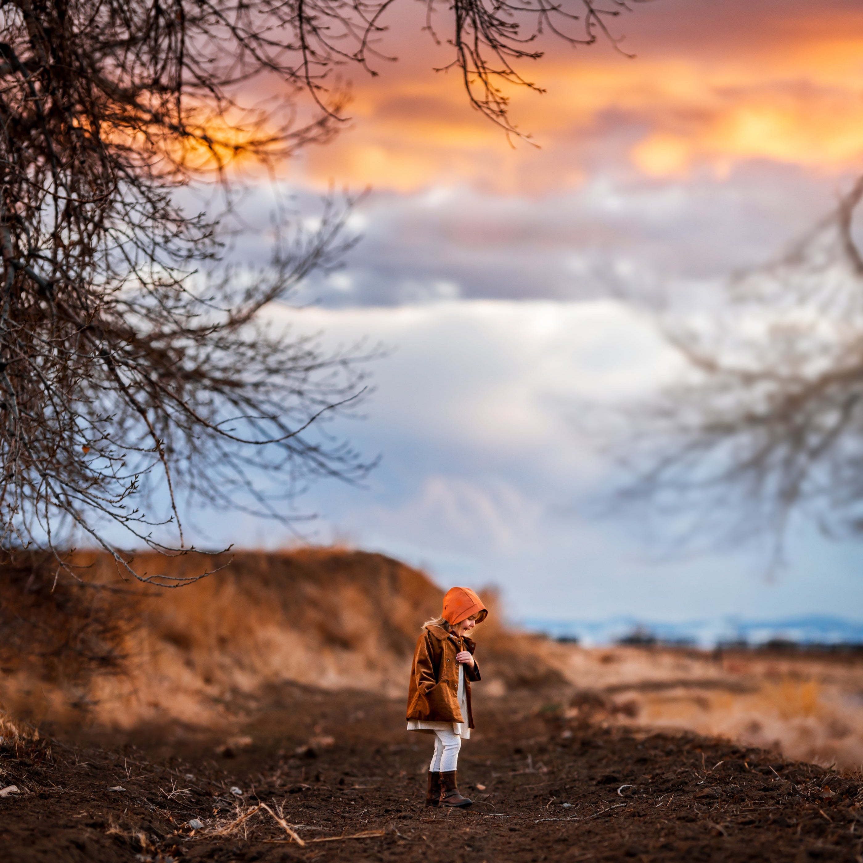 Child wearing a brown coat and bonnet against a blurred natural background with clouds