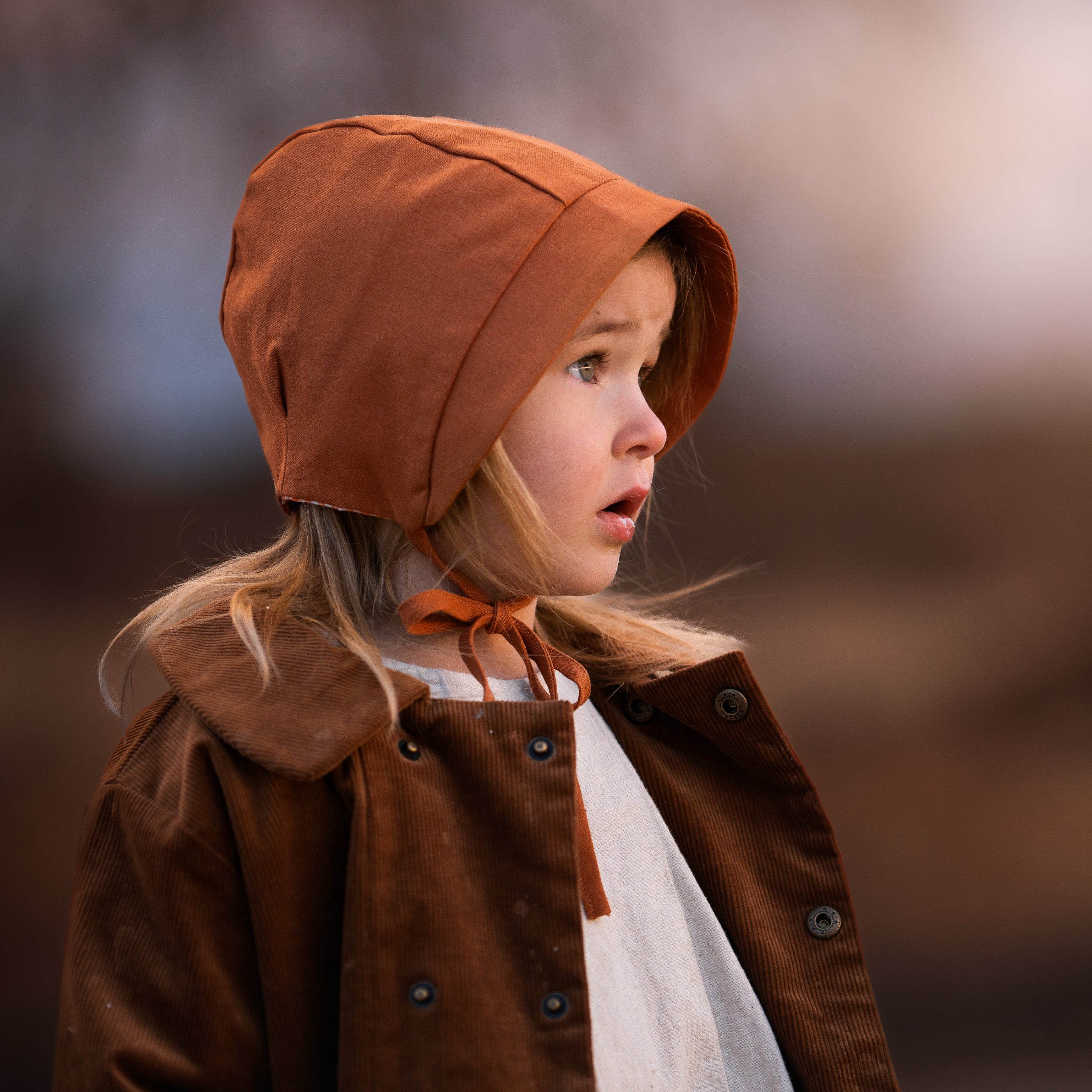Child wearing a brown coat and bonnet against a blurred natural background