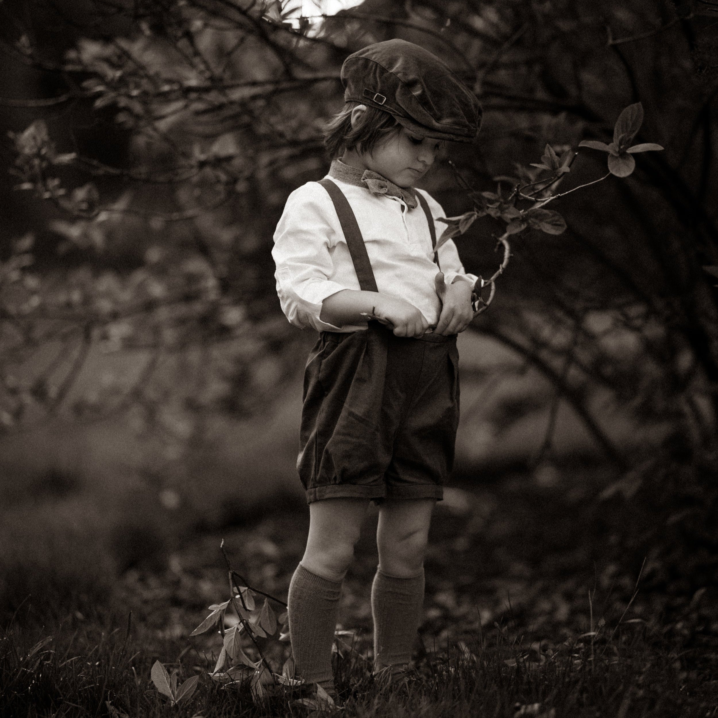 Black and white photo of a child in suspenders and shorts standing in a natural setting.