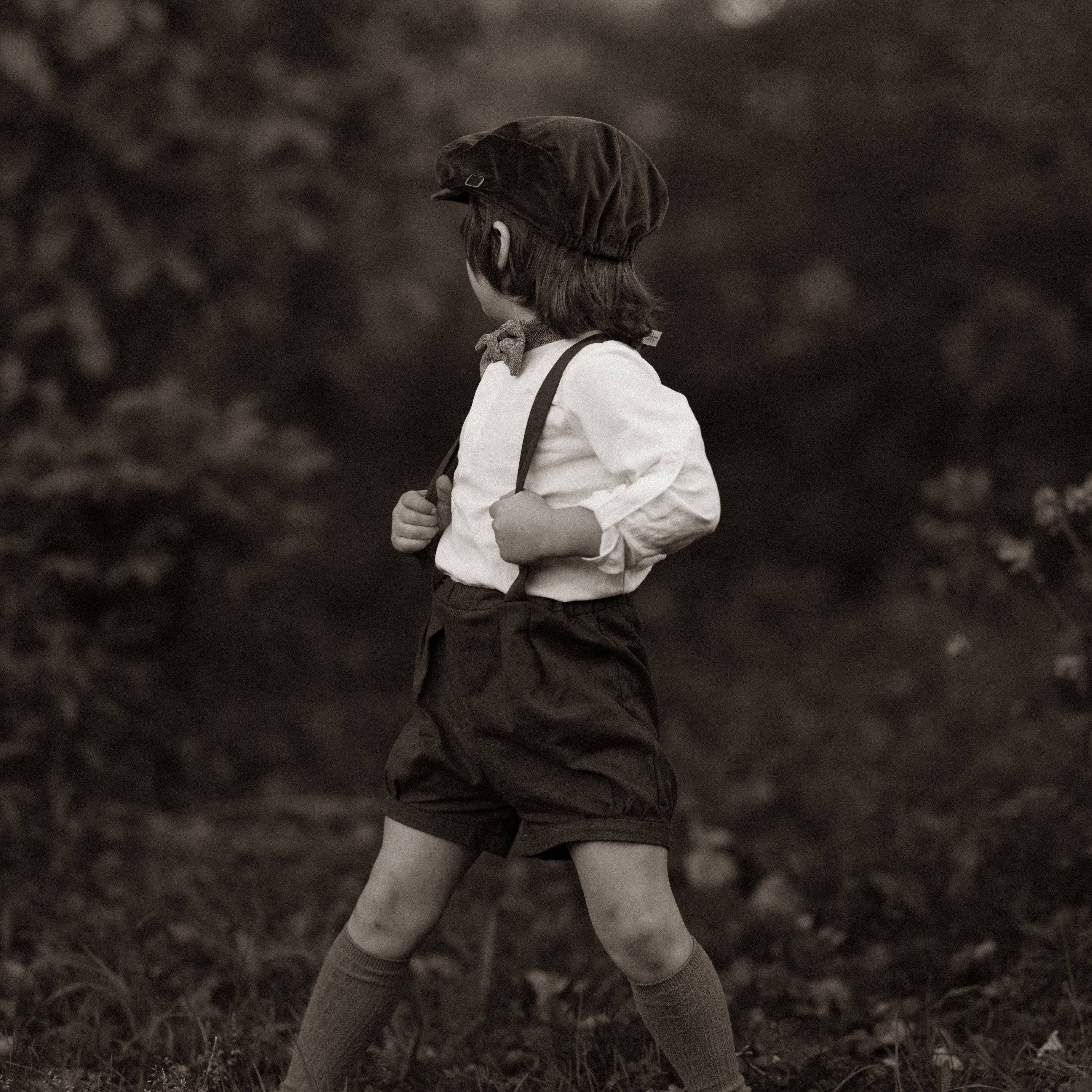 A model boy holding the adjustable straps of his black linen suspender shorts, emphasizing the customized fit and style, with a focus on his comfort and the details of his outfit in a natural setting.