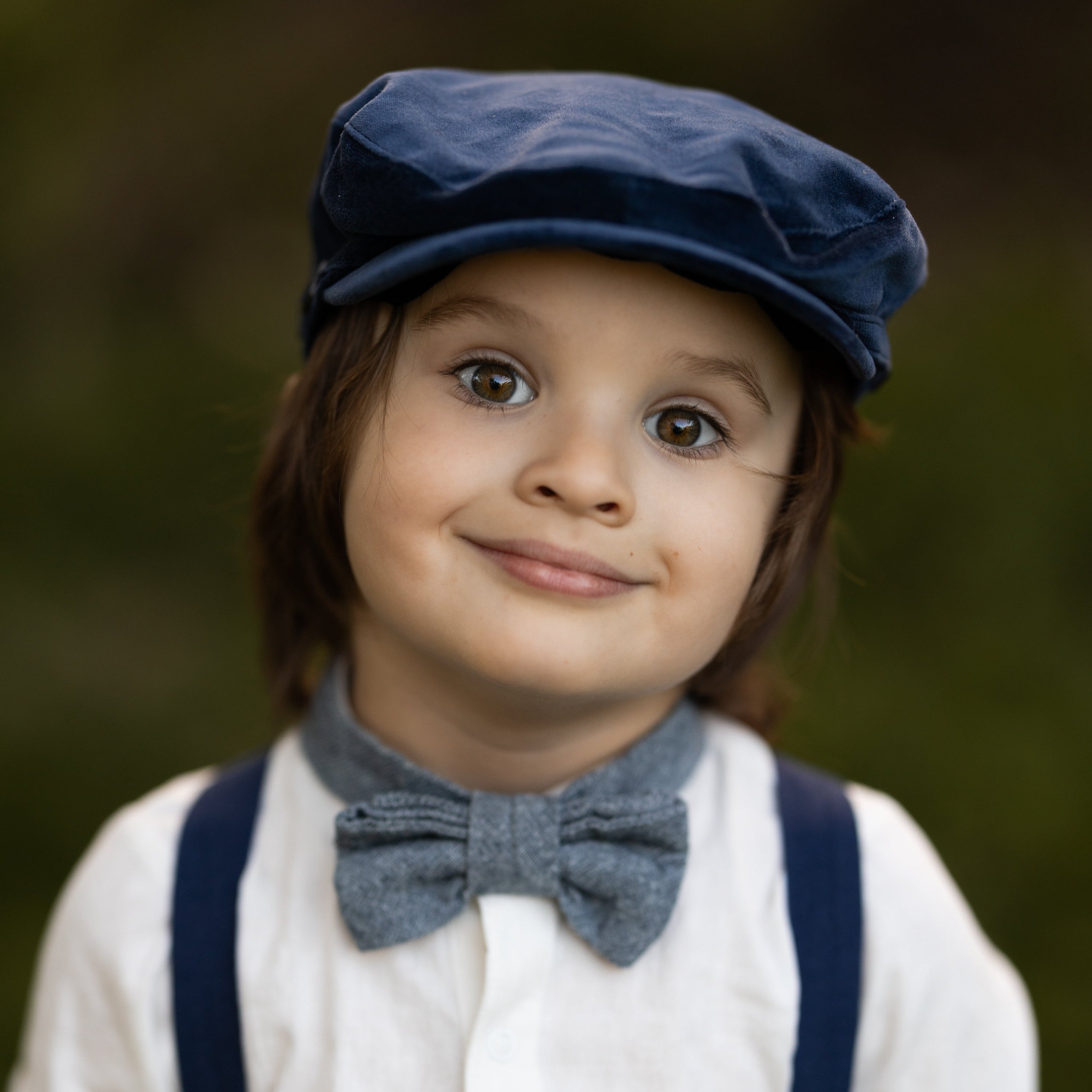 Child wearing a blue cap, suspenders, and bow tie against a blurred background