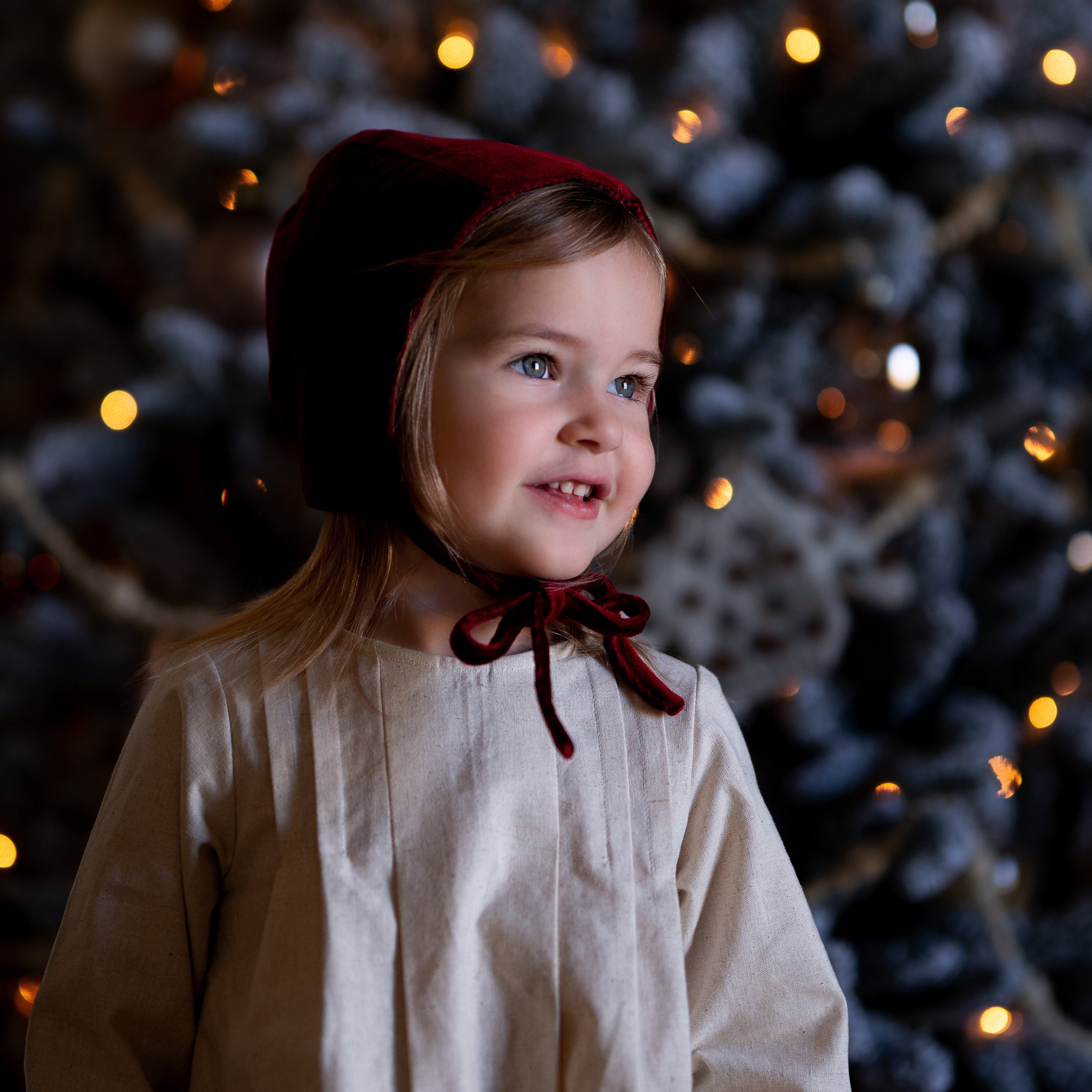 Child wearing a red bonnet in front of a decorated Christmas tree with lights.