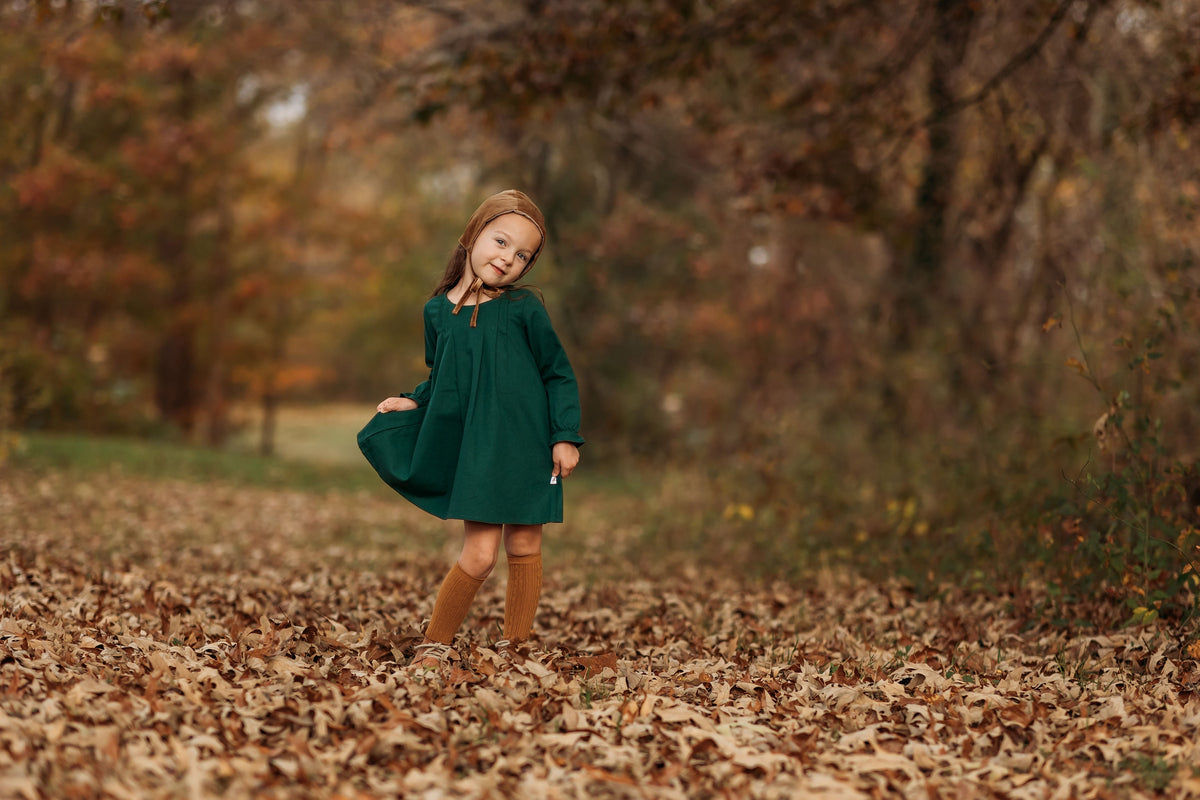A 4-year-old girl posing sweetly while holding the tip of her dress’s skirt, showcasing the stylish fit and comfortable design of the evergreen linen dress.