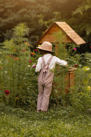 A young child wearing a beige hat, white shirt, and red-and-white gingham overalls stands in a lush garden beside a small wooden bee house, viewed from behind.