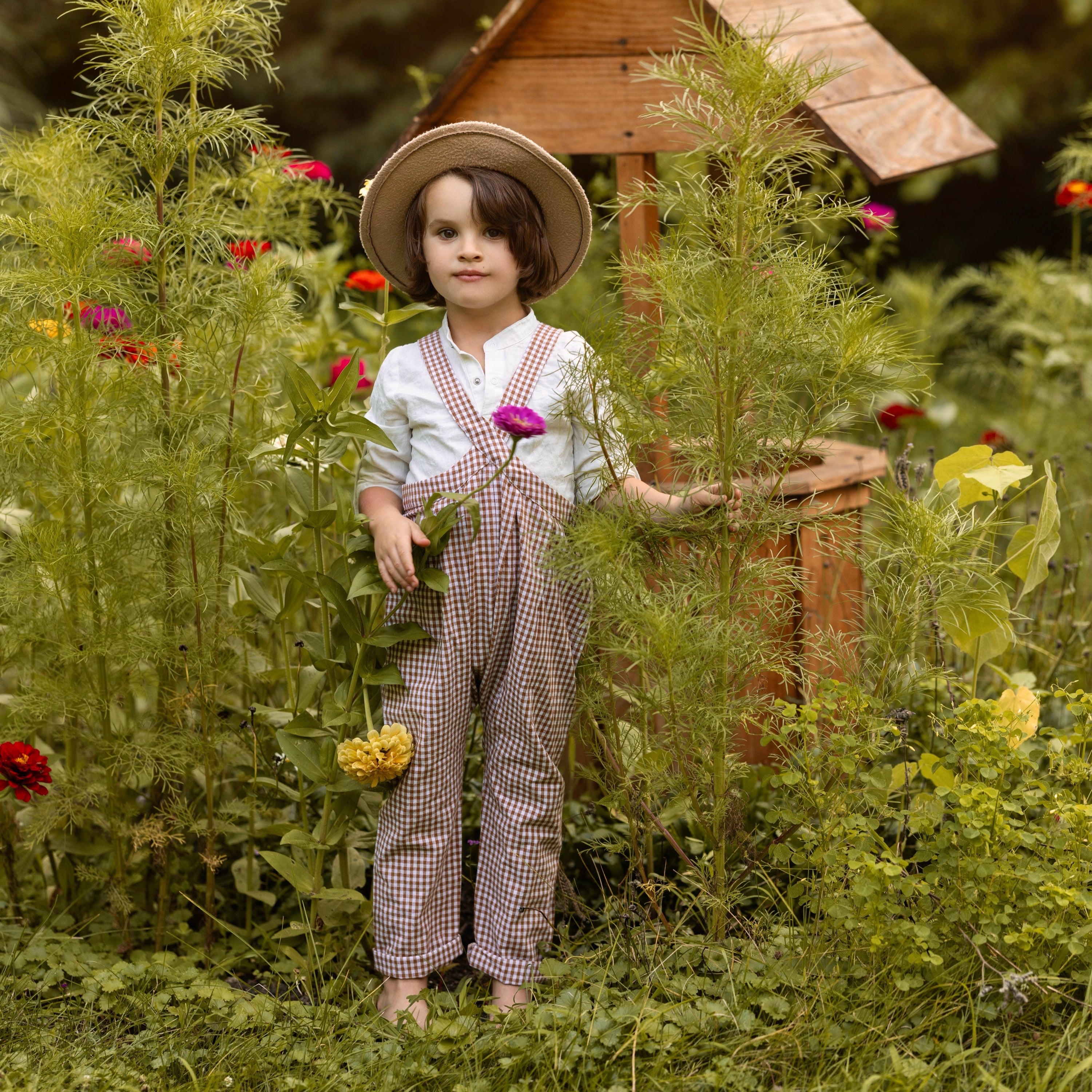 Child in a garden with a small wooden house and flowers dressed in a hat, white shirt and vintage-inspired overalls in brown gingham.