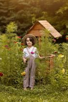 A young child stands barefoot in a lush garden beside a small wooden birdhouse, wearing a straw hat, white shirt, and brown-and-white gingham overalls while holding a pink flower.