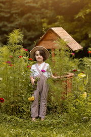 A young child stands barefoot in a lush garden beside a small wooden birdhouse, wearing a straw hat, white shirt, and brown-and-white gingham overalls while holding a pink flower.