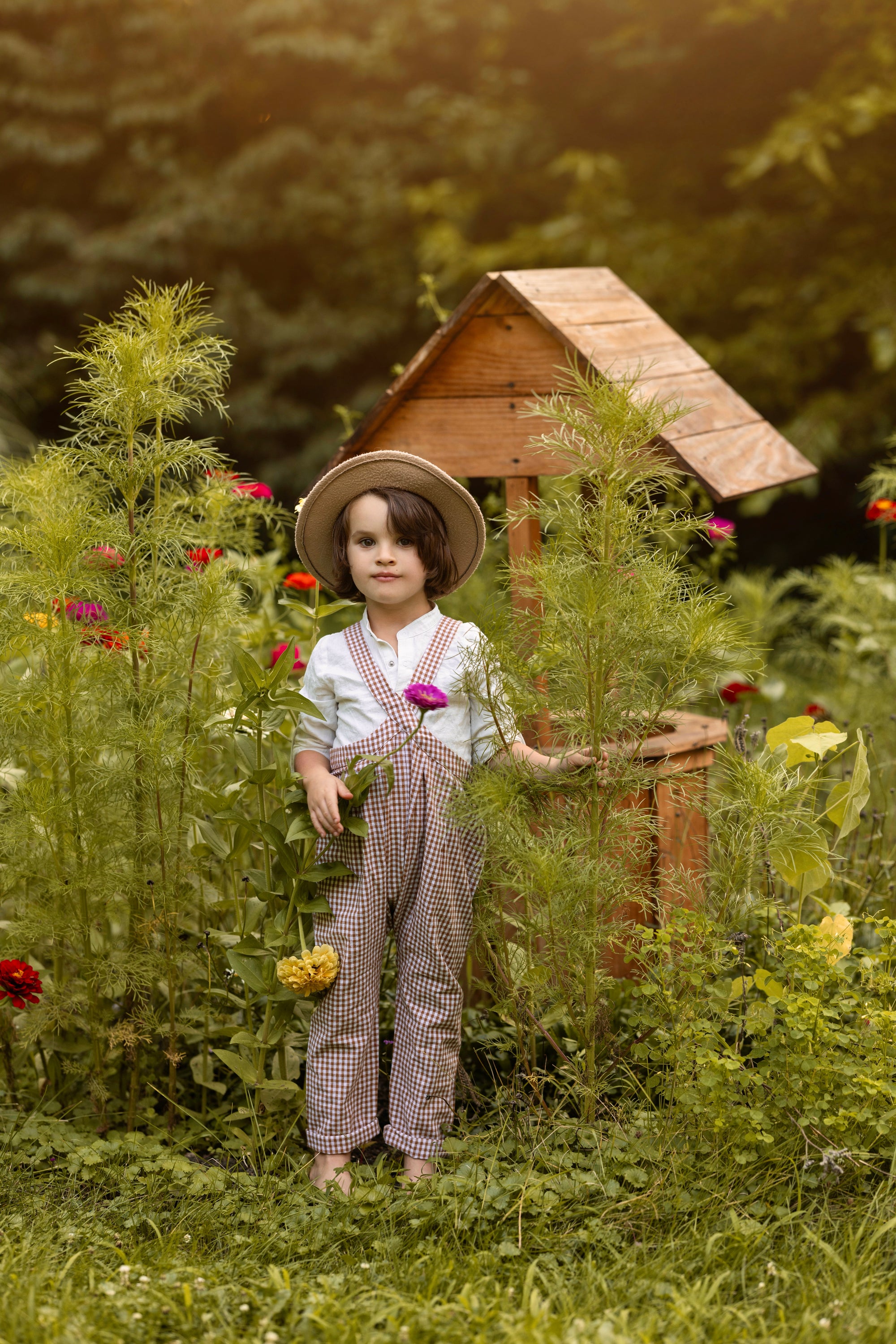A young child stands barefoot in a lush garden beside a small wooden birdhouse, wearing a straw hat, white shirt, and brown-and-white gingham overalls while holding a pink flower.