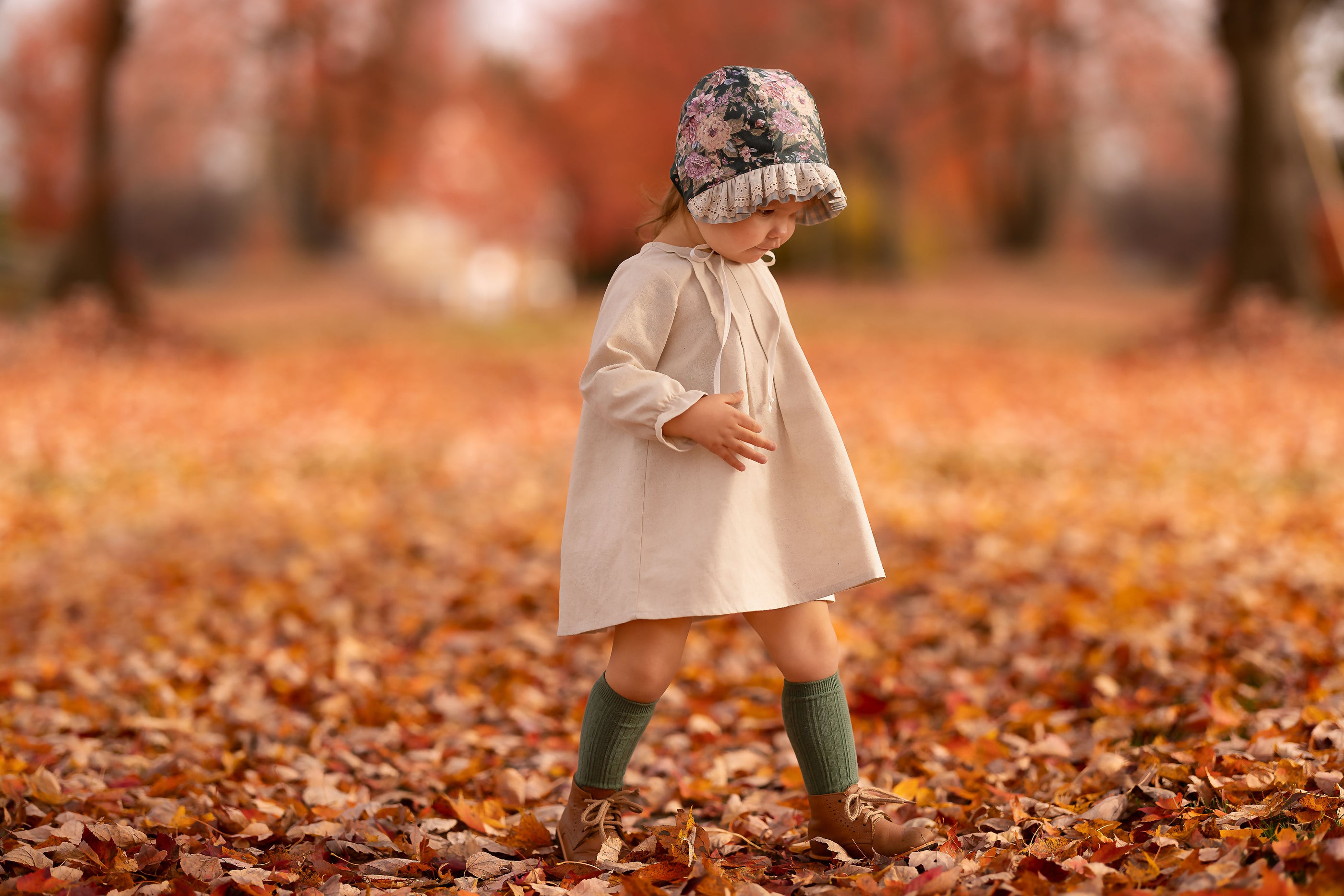 A toddler girl wearing a cream-colored dress, a floral knit beanie, green socks, and brown boots walks through a path covered in fallen autumn leaves in a park.