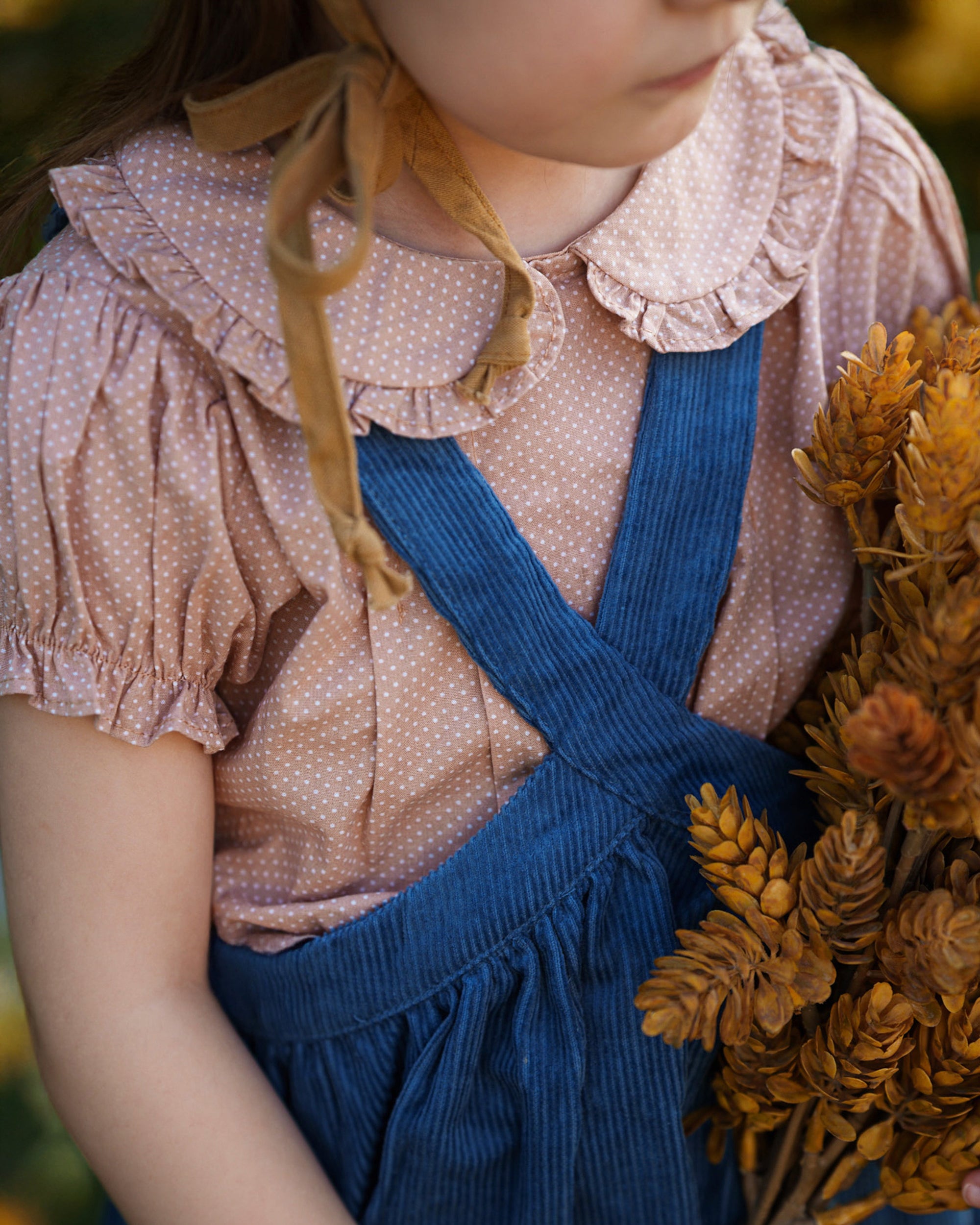 A close-up of a young child wearing a pink polka-dot blouse with a ruffled collar and tan ribbon tie, paired with blue corduroy overalls, holding a bouquet of dried golden wheat and pinecones.