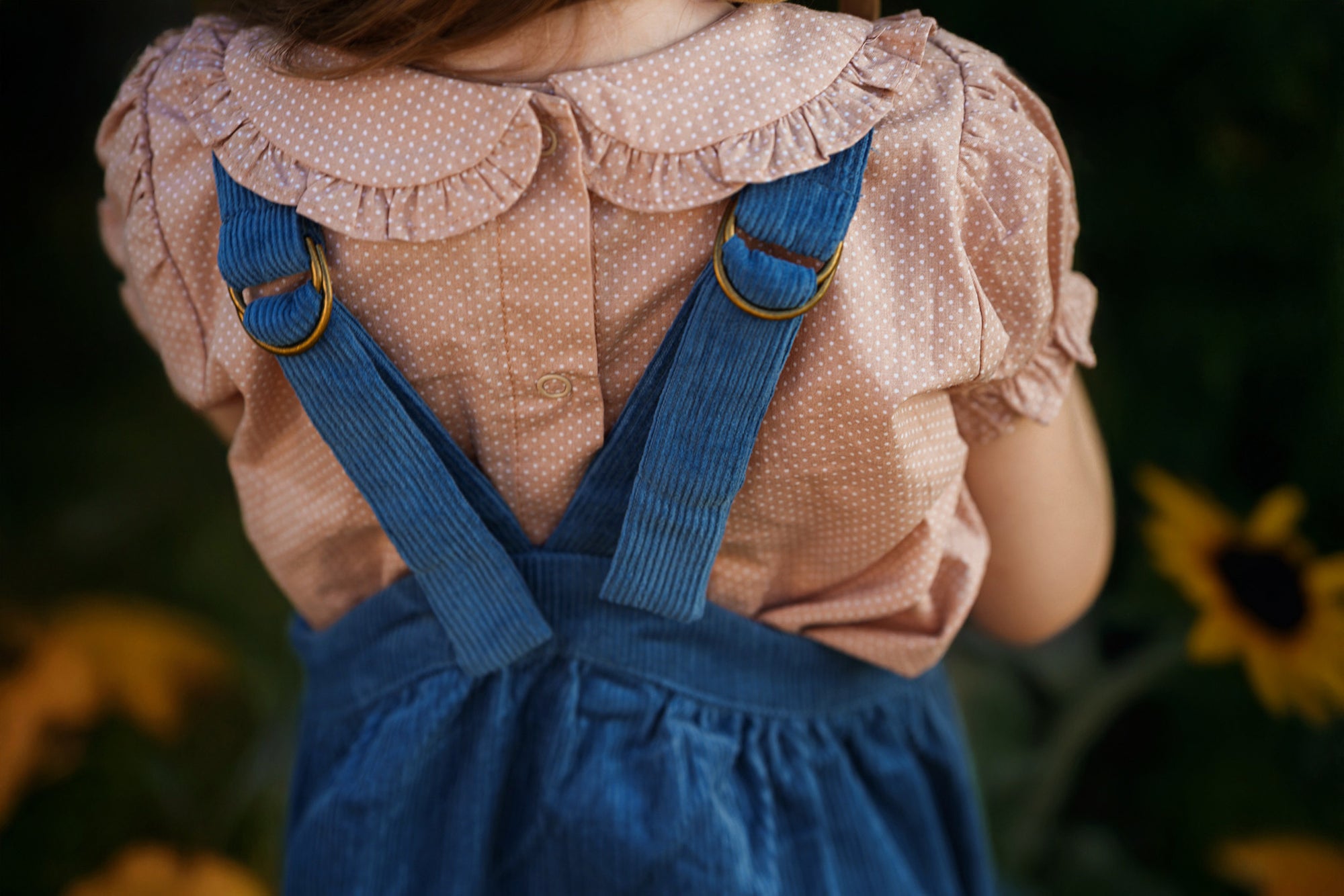 A child viewed from behind wearing a pink polka-dot blouse with a ruffled Peter Pan collar and blue corduroy-style overalls with brass-tone ring hardware, set against a dark outdoor background with blurred yellow flowers.