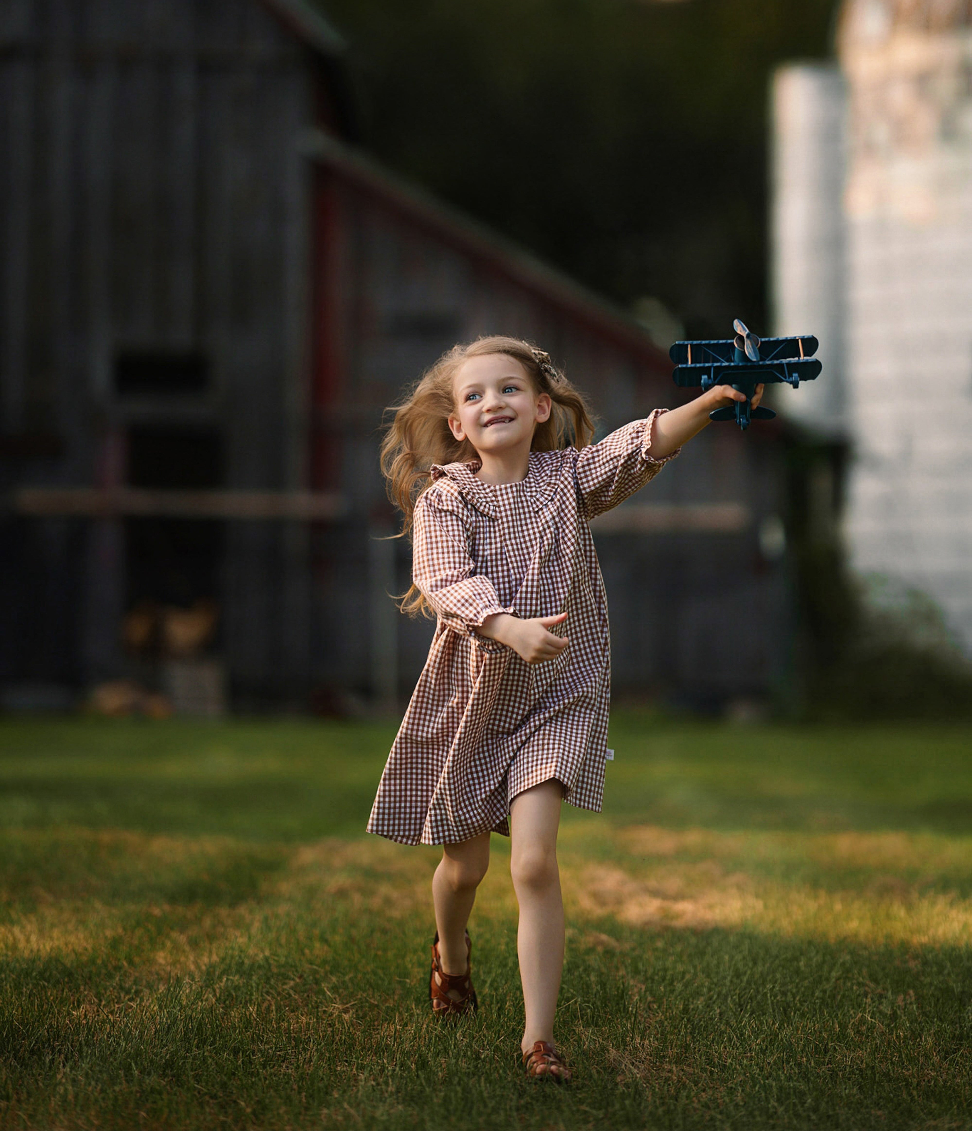 Young girl holding a toy airplane in a rural setting with a barn and greenery wearing a brown gingham dress.