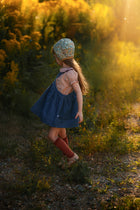 A young girl wearing a blue denim overall dress, a patterned knit bonnet, and rust-red knee-high socks stands in a sunlit outdoor field with golden wildflowers.