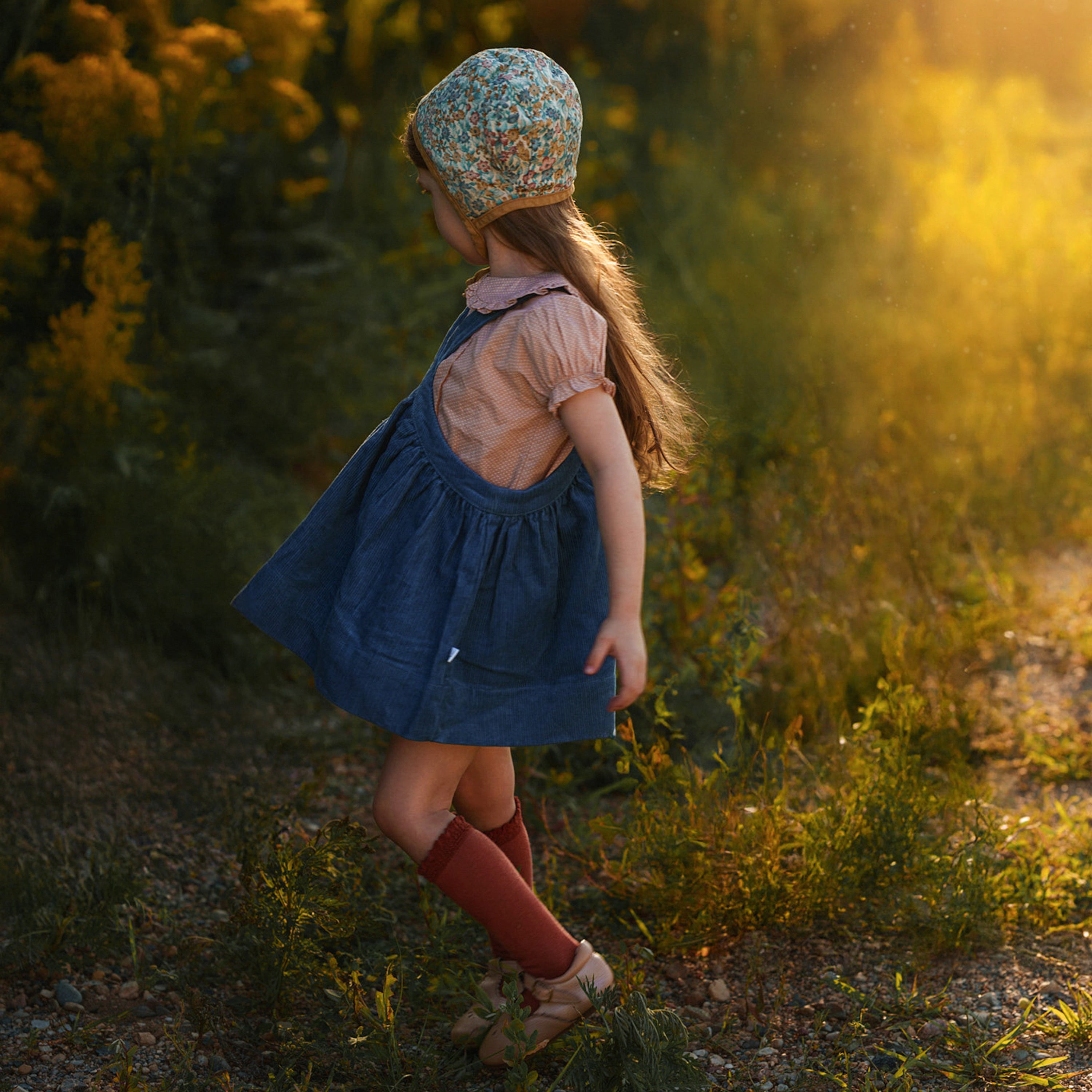 Young girl in a blue dress and floral hat walking through a sunlit forest