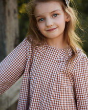 A young girl with blonde hair and blue eyes wearing a brown and white gingham long-sleeve dress, photographed outdoors against a soft-focus background.