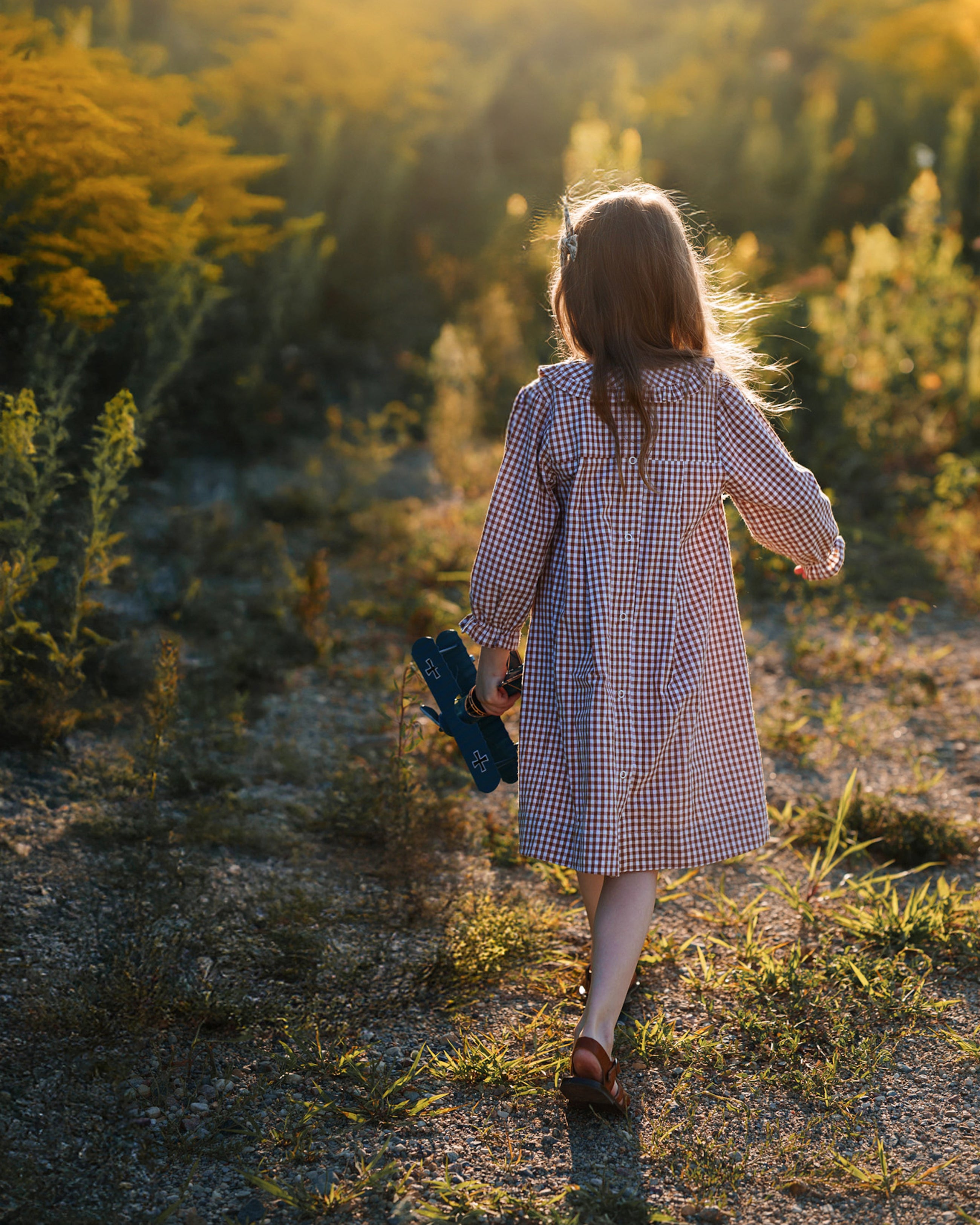 A young girl seen from behind walking on a sunlit path through a grassy field, wearing a red and white gingham dress and holding a pair of dark blue shoes.
