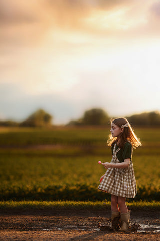 Young girl standing in a field with a sunset in the background in a checkered pinafore eating a lollipop
