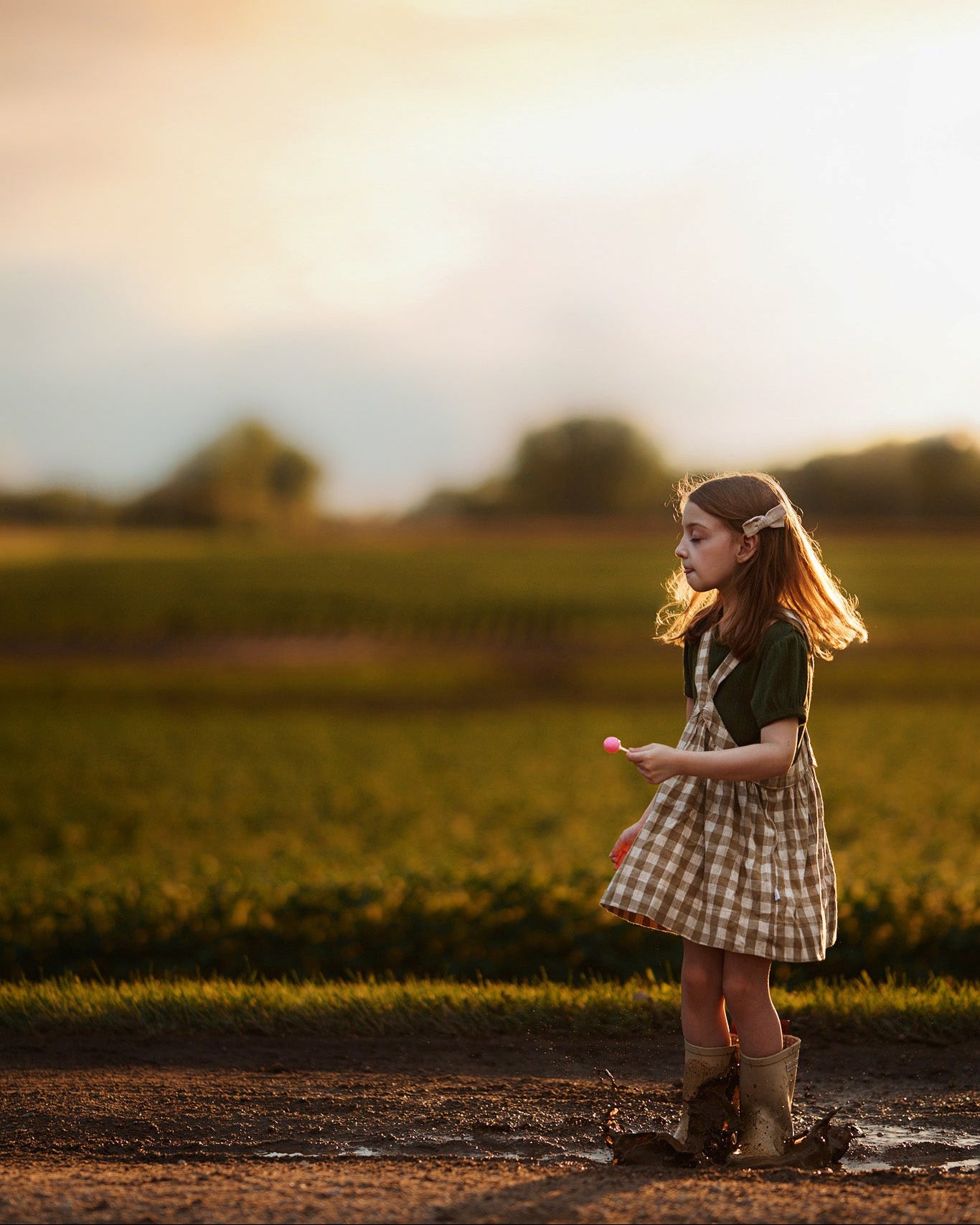 A young girl stands in a muddy field at sunset, wearing a brown and beige plaid pinafore dress over a dark green shirt and brown boots.
