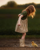 A young girl with red hair stands in a muddy puddle wearing a green short-sleeve top and a beige-and-white gingham pinafore dress, with beige rain boots. A small yellow duck is standing nearby in the mud on a dirt path, with a blurred green field and sunset sky in the background.