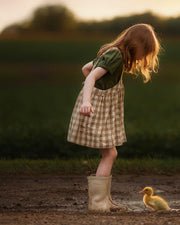 A young girl with red hair stands in a muddy puddle wearing a green short-sleeve top and a beige-and-white gingham pinafore dress, with beige rain boots. A small yellow duck is standing nearby in the mud on a dirt path, with a blurred green field and sunset sky in the background.