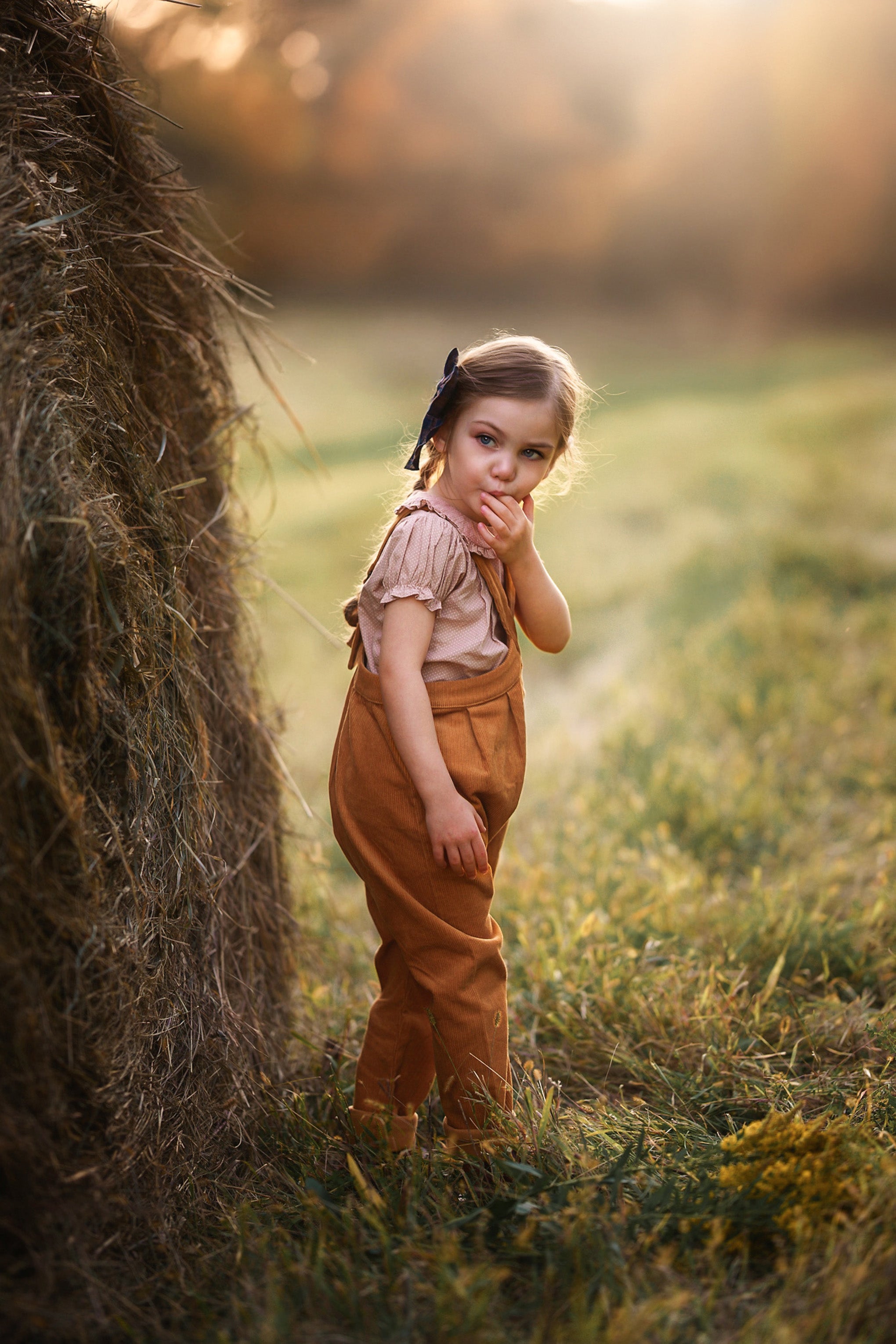 A young child standing outdoors beside a large hay bale in a grassy field, wearing a light pink top and brown overalls, with warm golden sunlight and a blurred background.
