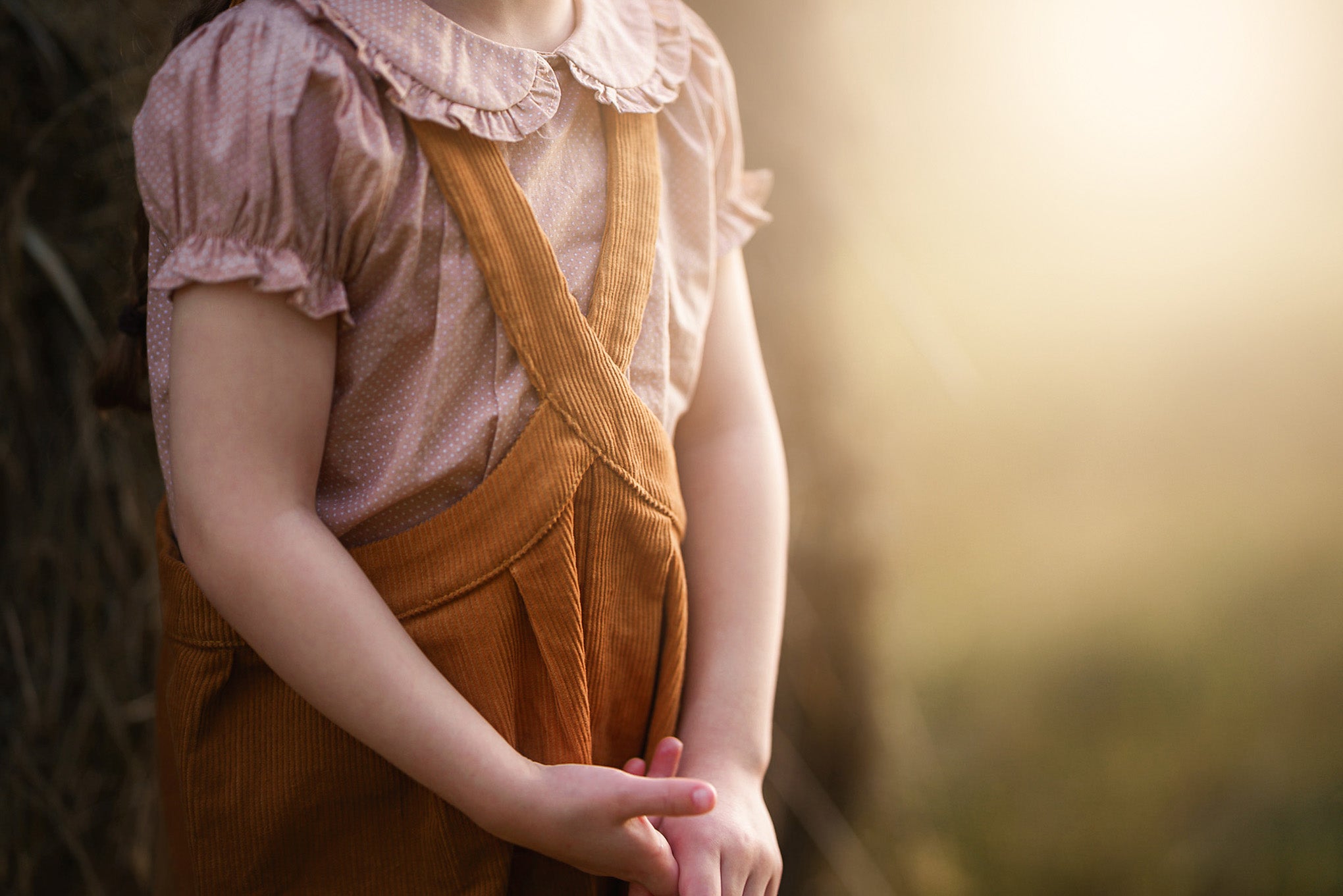 A child wearing a pink ruffled blouse and brown corduroy overalls, standing outdoors in warm golden light with a blurred natural background.