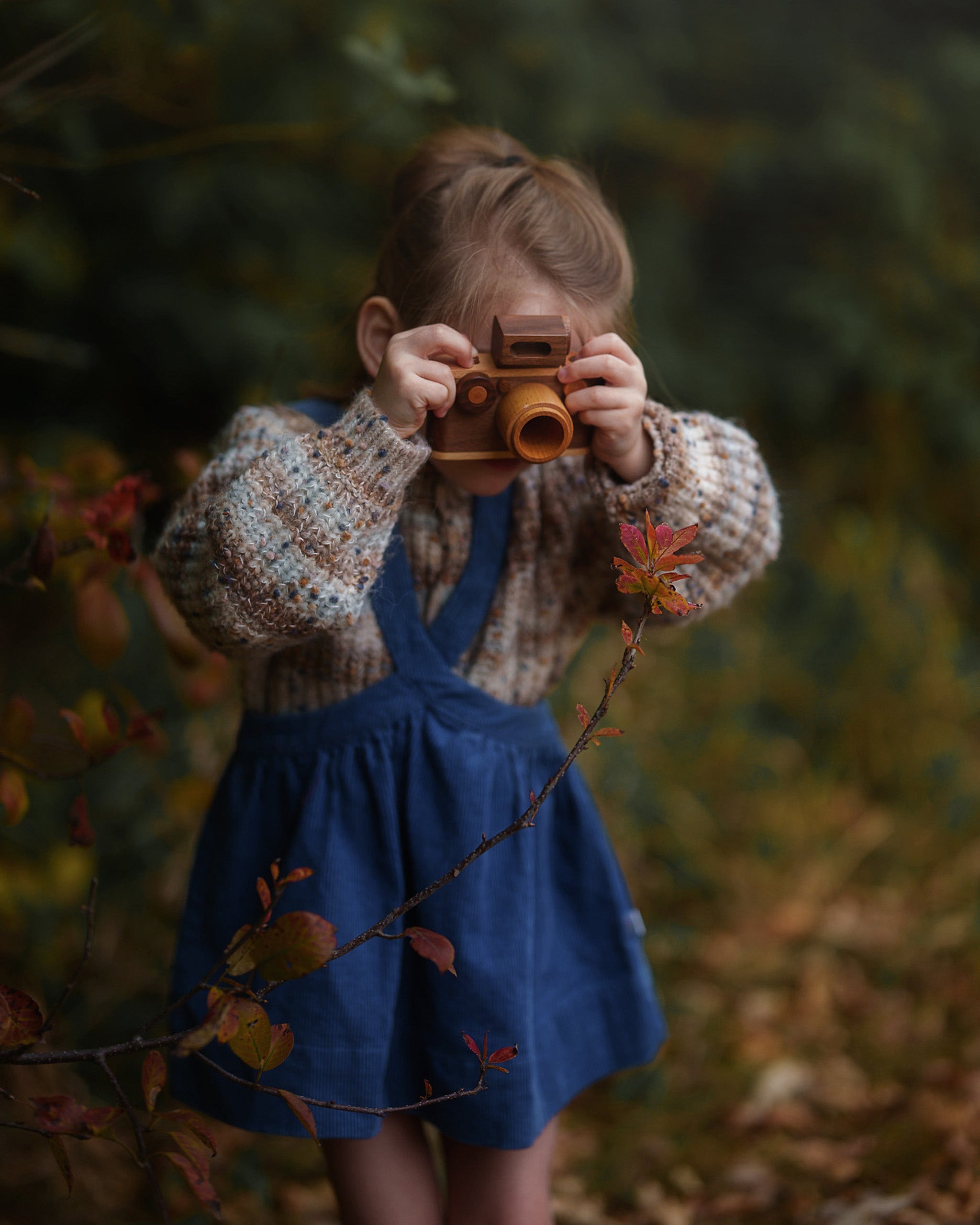 Little girl holding a vintage camera in a forest setting wearing a blue pinafore dress over a chunky sweater.