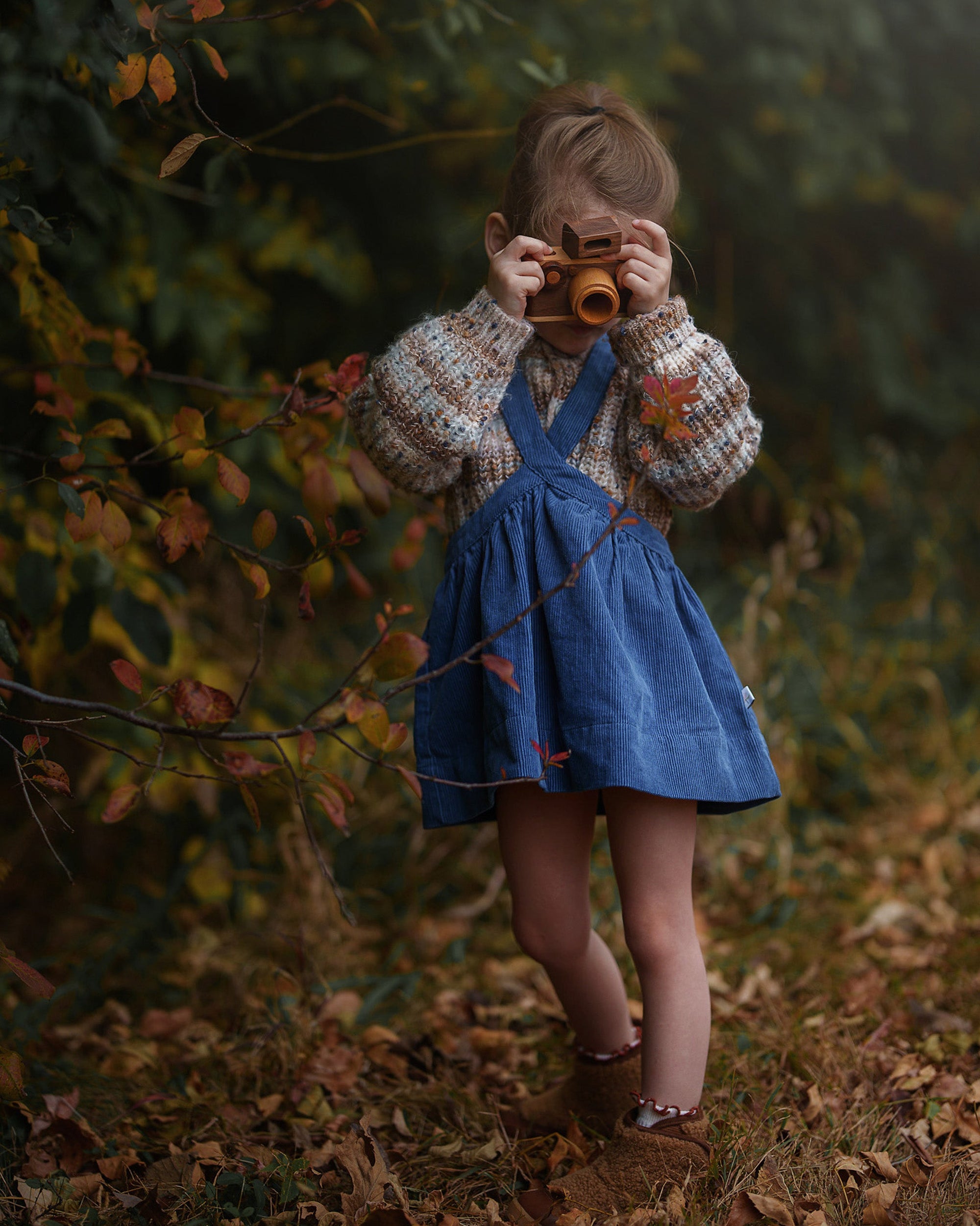A young child wearing a blue denim pinafore dress over a cream and brown knit sweater stands in a leafy autumn forest holding a vintage-style camera to their face.