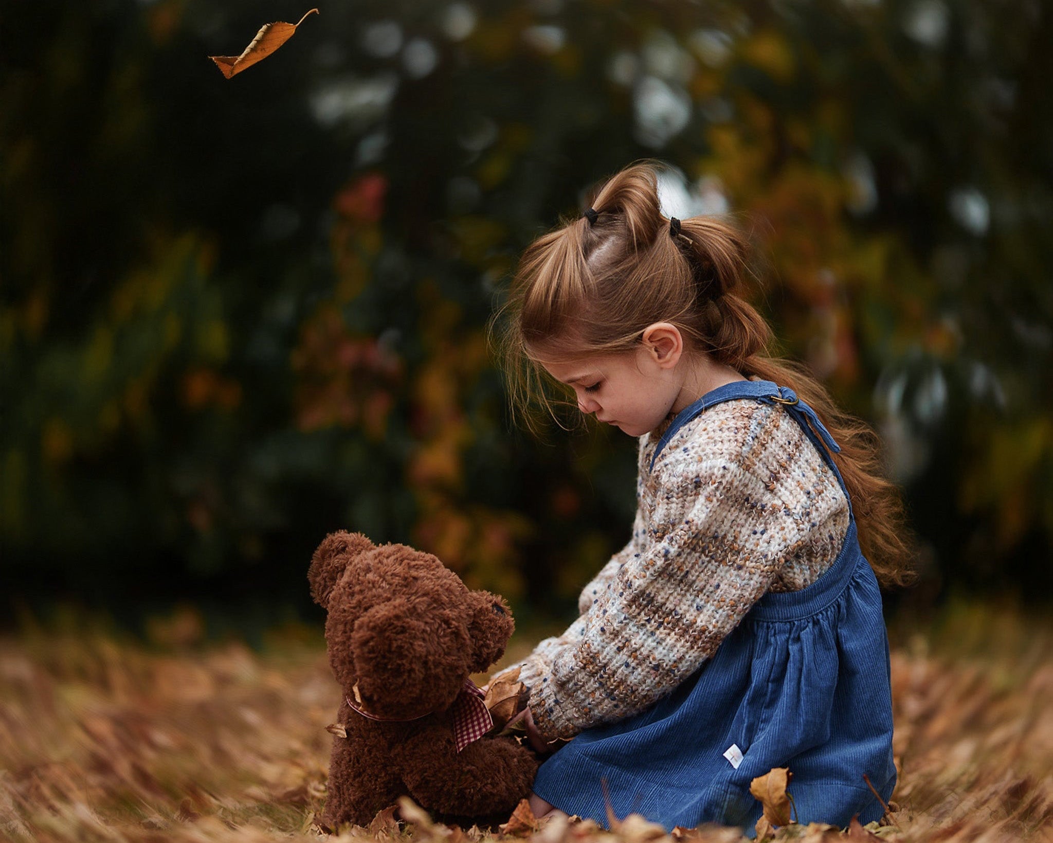 Child holding a teddy bear in an outdoor setting with trees and leaves in a blue corduroy pinafore.