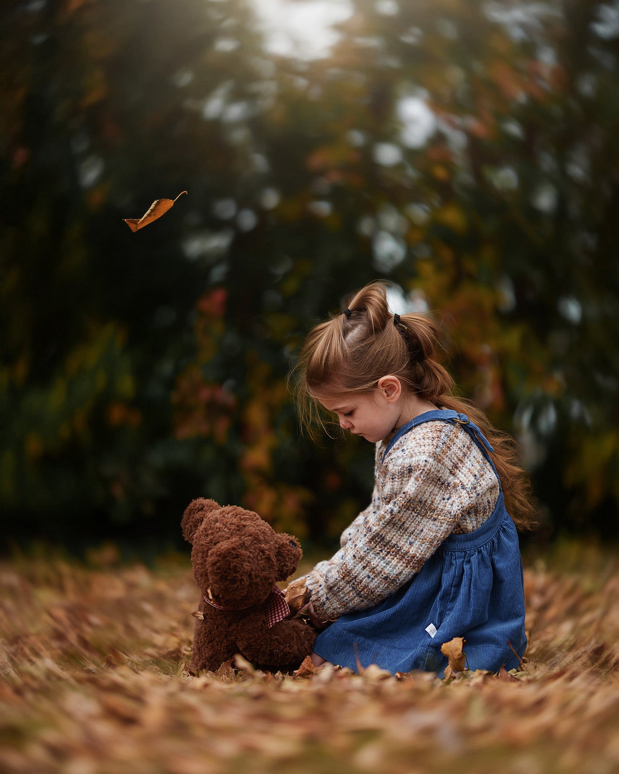A young girl in a brown plaid shirt and blue denim overalls sits on a bed of fallen leaves in an autumn park, holding a brown teddy bear.