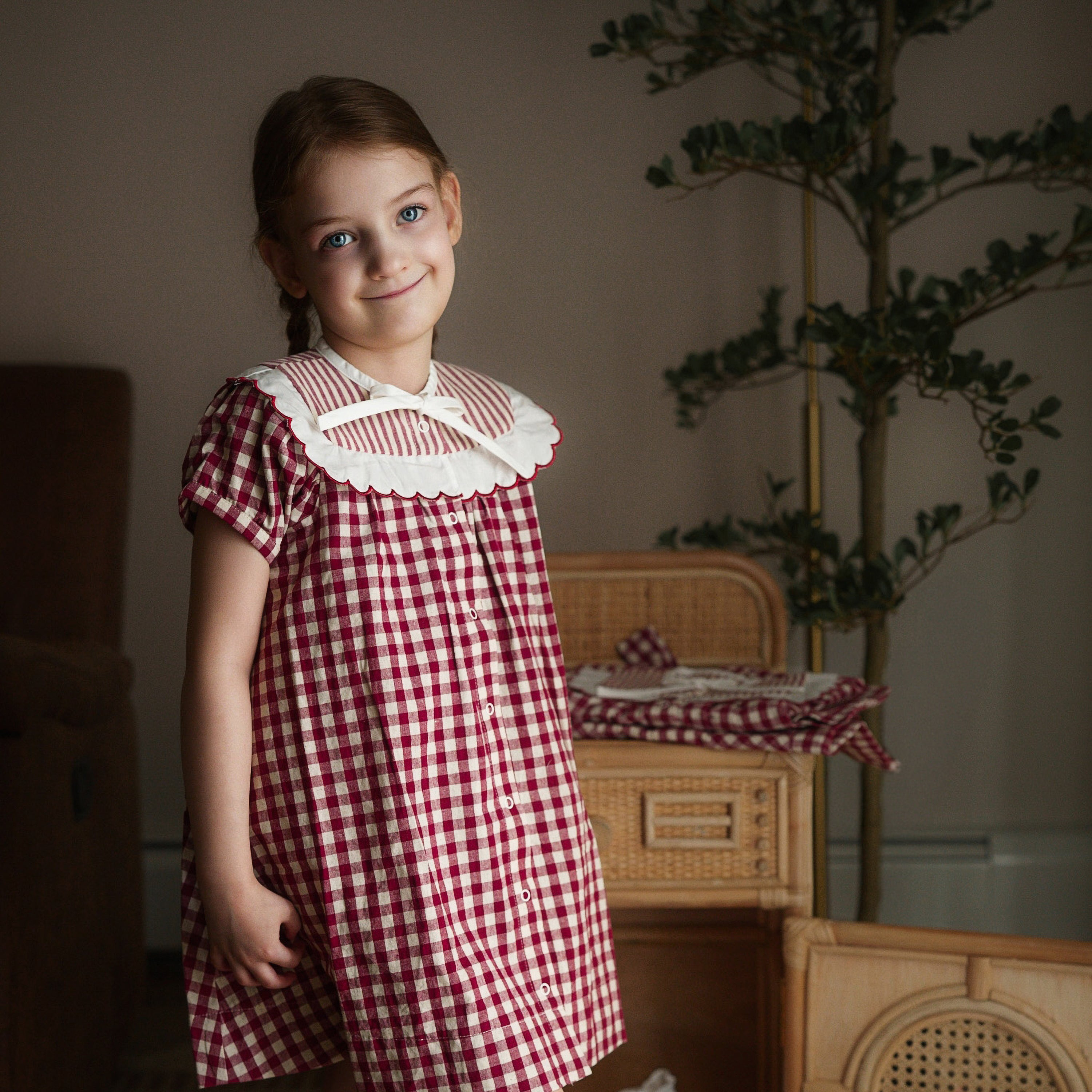 Young girl in a red and white checkered dress standing in a room with wicker furniture and plants.
