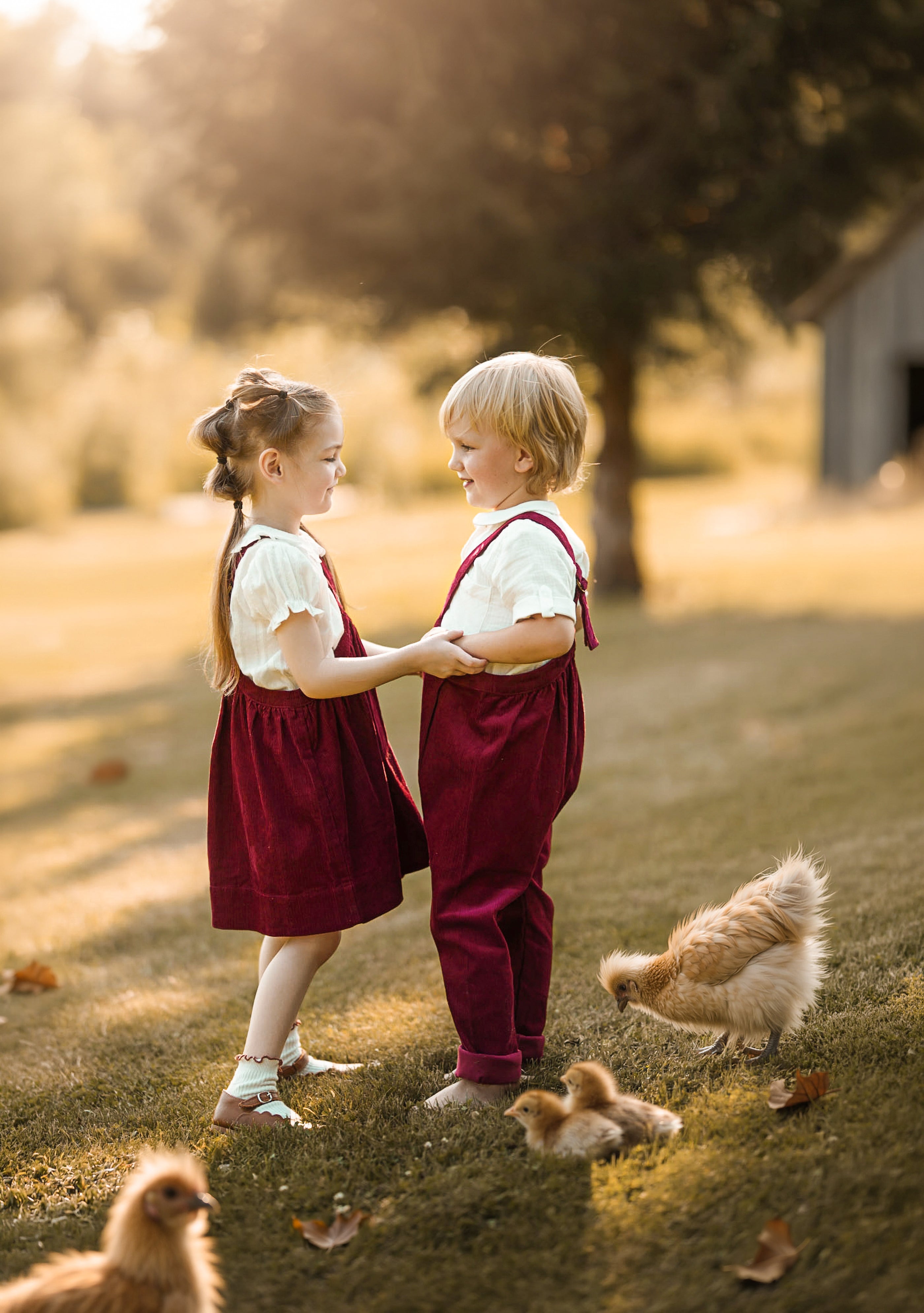 Two young children standing on a grassy lawn outdoors, wearing matching burgundy pinafore-style dresses over white shirts. Chickens are visible around them in a sunlit yard with trees in the background.