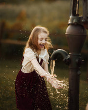 A young girl with long light brown hair plays with water from a vintage metal hand pump in an outdoor garden setting during golden hour. She is wearing a cream-colored blouse and a dark burgundy velvet pinafore dress, with water droplets splashing around her.