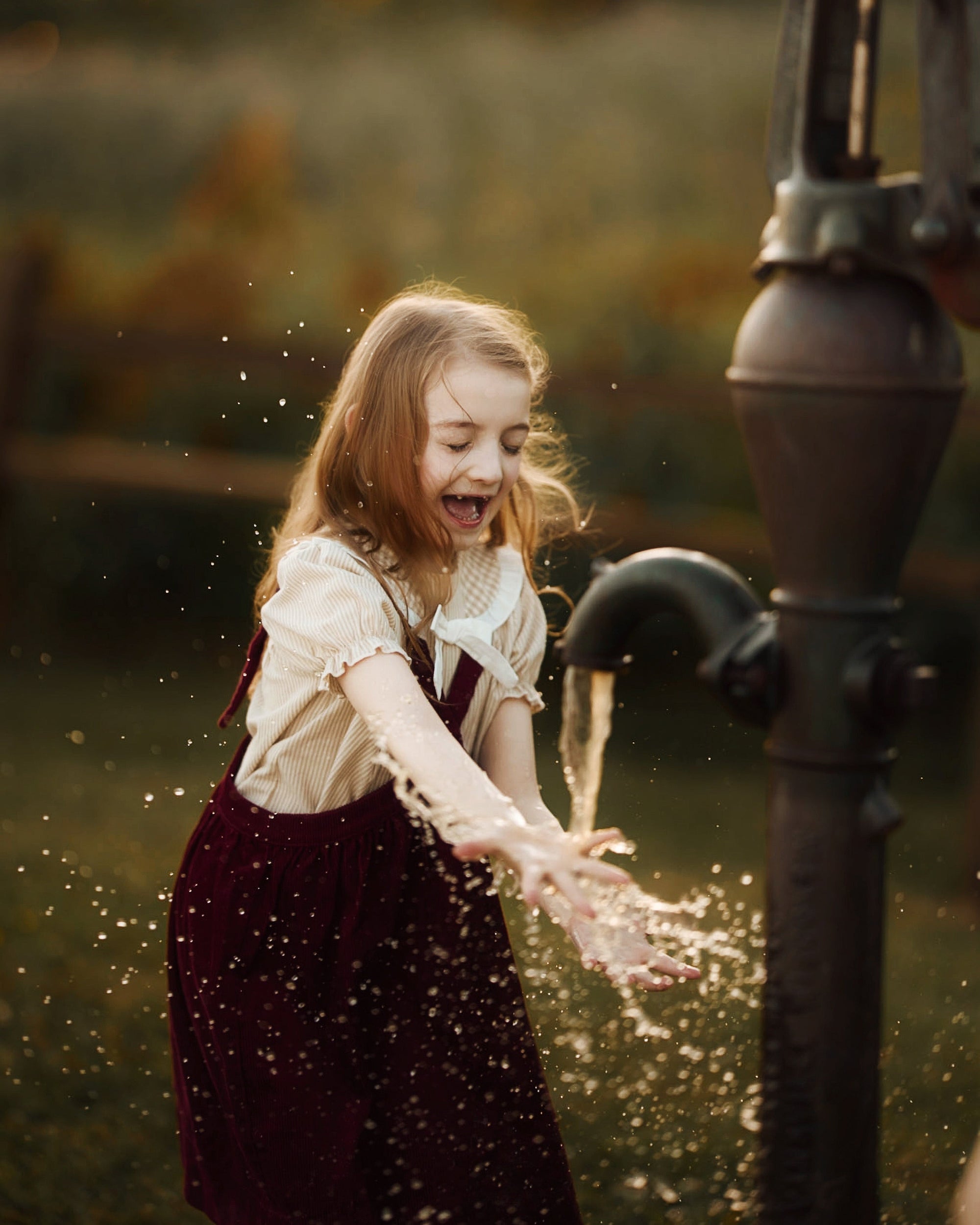 A young girl with long light brown hair plays with water from a vintage metal hand pump in an outdoor garden setting during golden hour. She is wearing a cream-colored blouse and a dark burgundy velvet pinafore dress, with water droplets splashing around her.