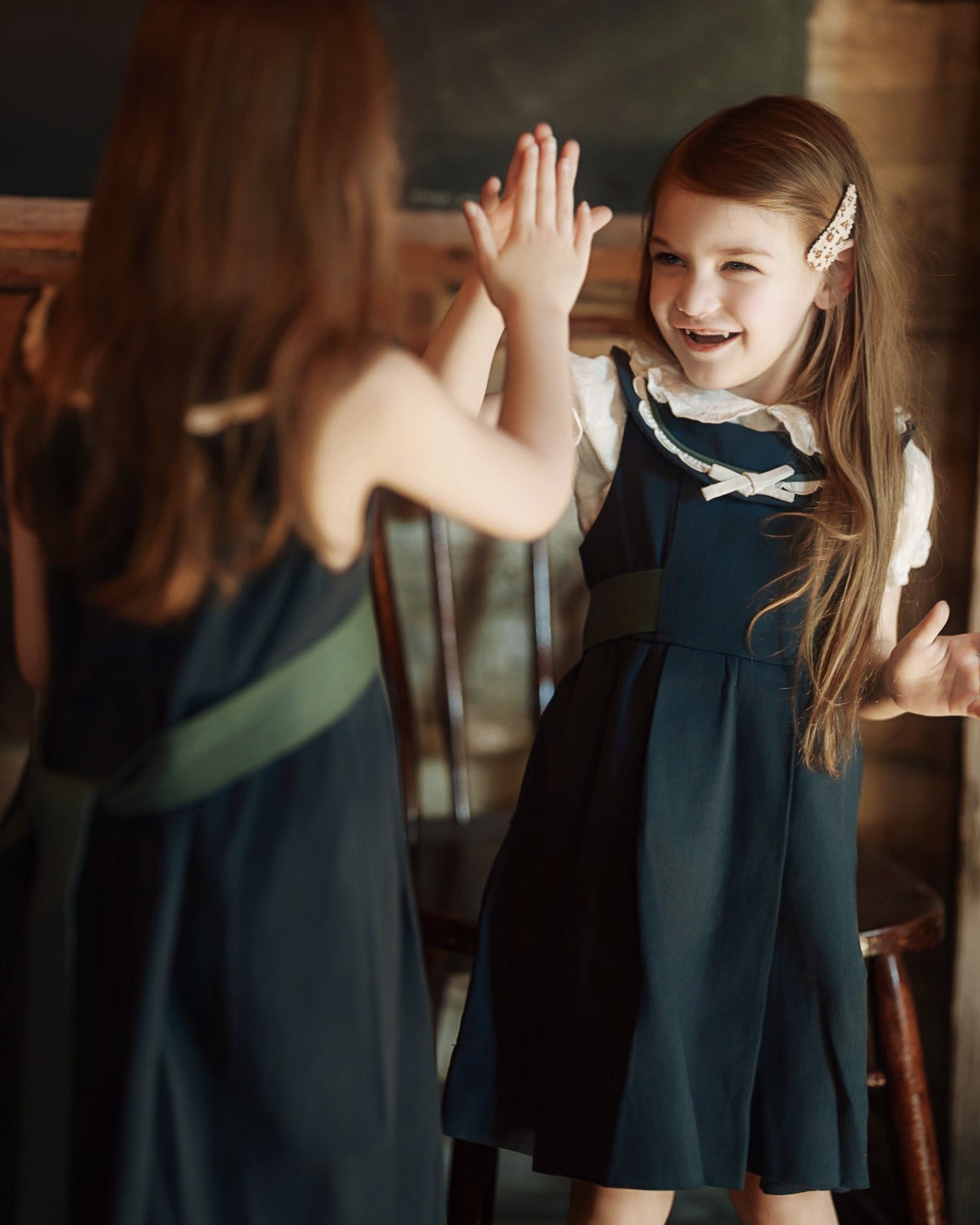 Two young girls in matching navy blue dresses with white collars and sashes pose for a high-five in a rustic indoor setting with warm, dim lighting.