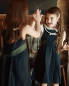 Two young girls in matching navy blue dresses with white collars and sashes pose for a high-five in a rustic indoor setting with warm, dim lighting.
